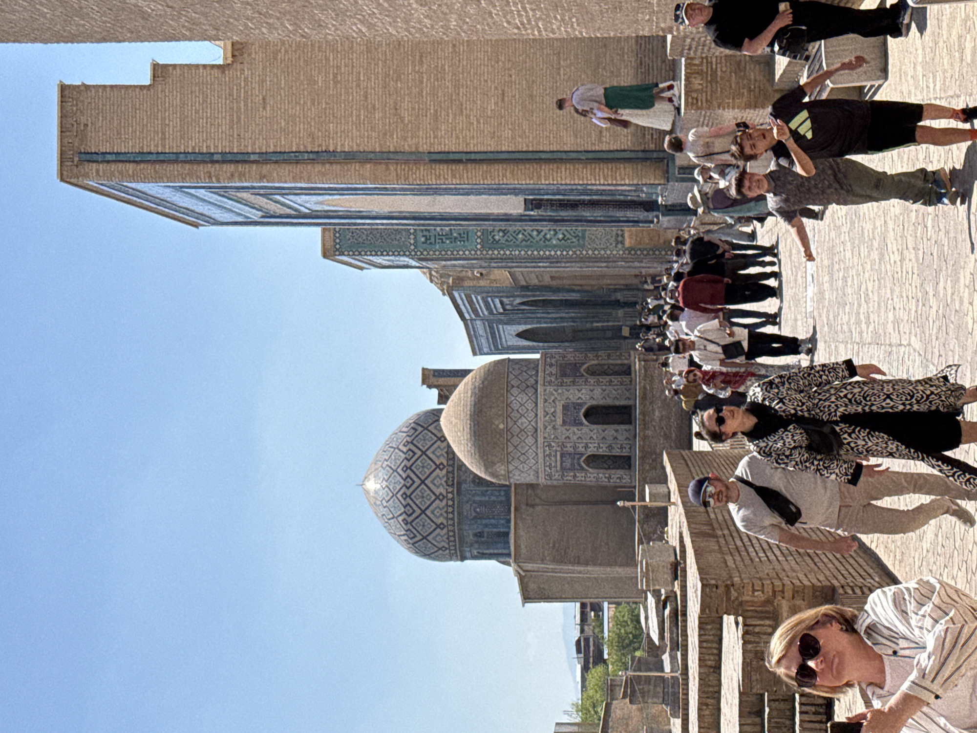 The image captures a bright, sunny day at the Shah-i-Zinda necropolis in Samarkand City, Uzbekistan. The scene presents a multitude of tourists milling about the stone-paved area, likely exploring the historical site.
Architecturally, the frame includes distinct segments of several buildings. Intricate blue-tiled domes and walls are apparent, showcasing the decorative mosaic patterns typical of Islamic architecture.
The tourists are diverse in age and appearance, each dressed in casual, warm-weather attire. They walk, pose for photos, and interact with each other, immersed in the location's historical significance.
The sky is clear and blue, suggesting a dry, sunny climate typical of Uzbekistan.