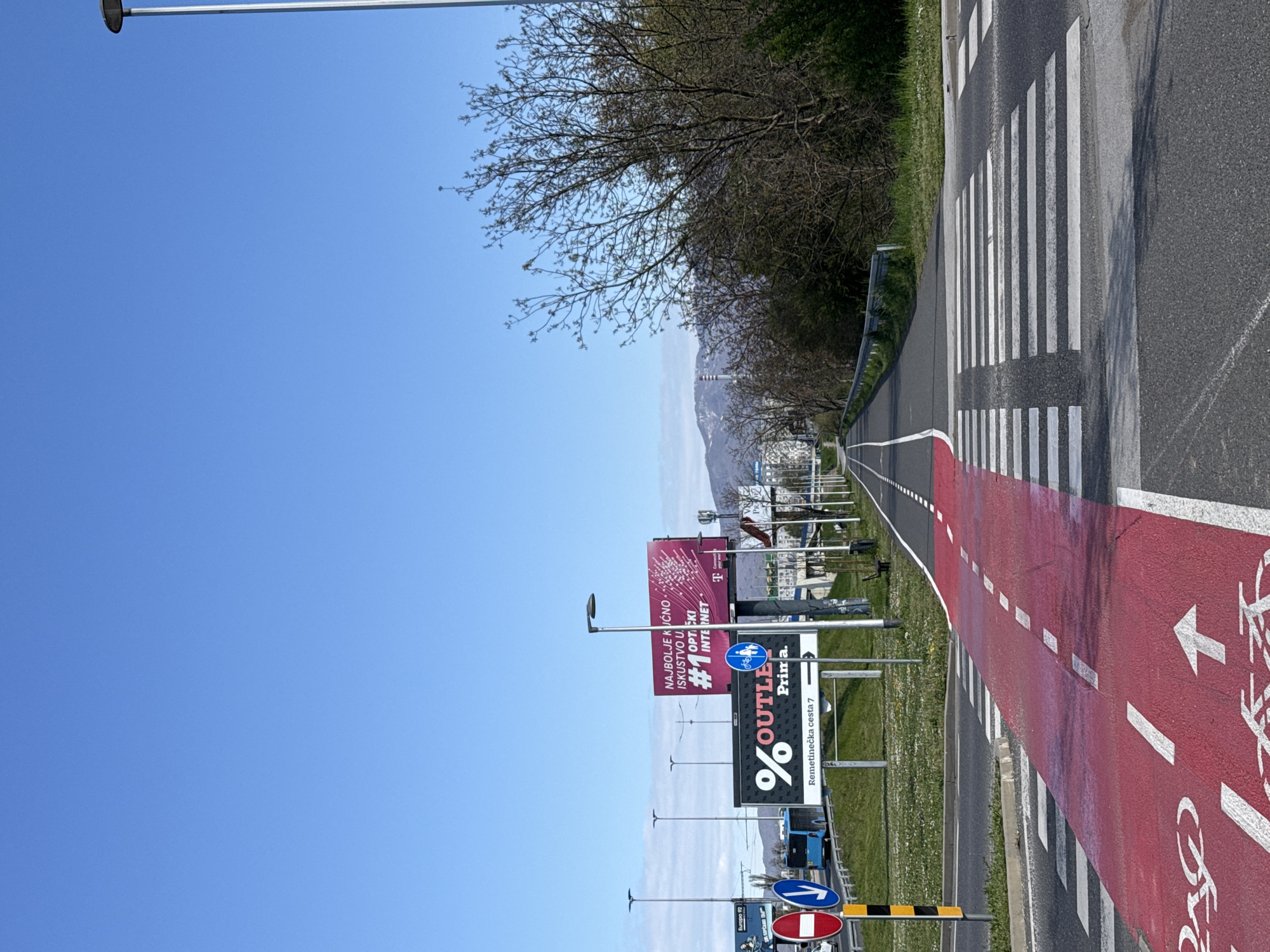 Here is a detailed description of the image:
**Scene Overview:** The image depicts an outdoor scene, predominantly featuring a bicycle path and a road. The shot is taken in daylight under a clear blue sky.
**Bicycle Path and Road:** The right side of the image shows a red-colored bicycle path with white lane markers and a bicycle symbol. Adjacent to it is an asphalt road marked with a pedestrian crosswalk.
**Advertising and Signage:** Along the left side, various advertising billboards and signs are visible, including promotional content for "% OUTLE Prima," a telecommunications company (likely T), and an advertisement highlighting the "#1 OPTICKI INTERNET." A street sign mentions "Remetinečka cesta 7". There is another sign that has a blue circle and a bicycle.
**Environmental Details:** A line of trees is located along the side of the road. In the background, a mountain range is visible.
**Location and Context:** Given the street sign mentioning "Remetinečka cesta," the location can be identified as being in the city of Zagreb, Croatia.
**Other Details:** There are utility poles with streetlights along the side of the road. A grassy area is seen between the road and the line of advertisements.