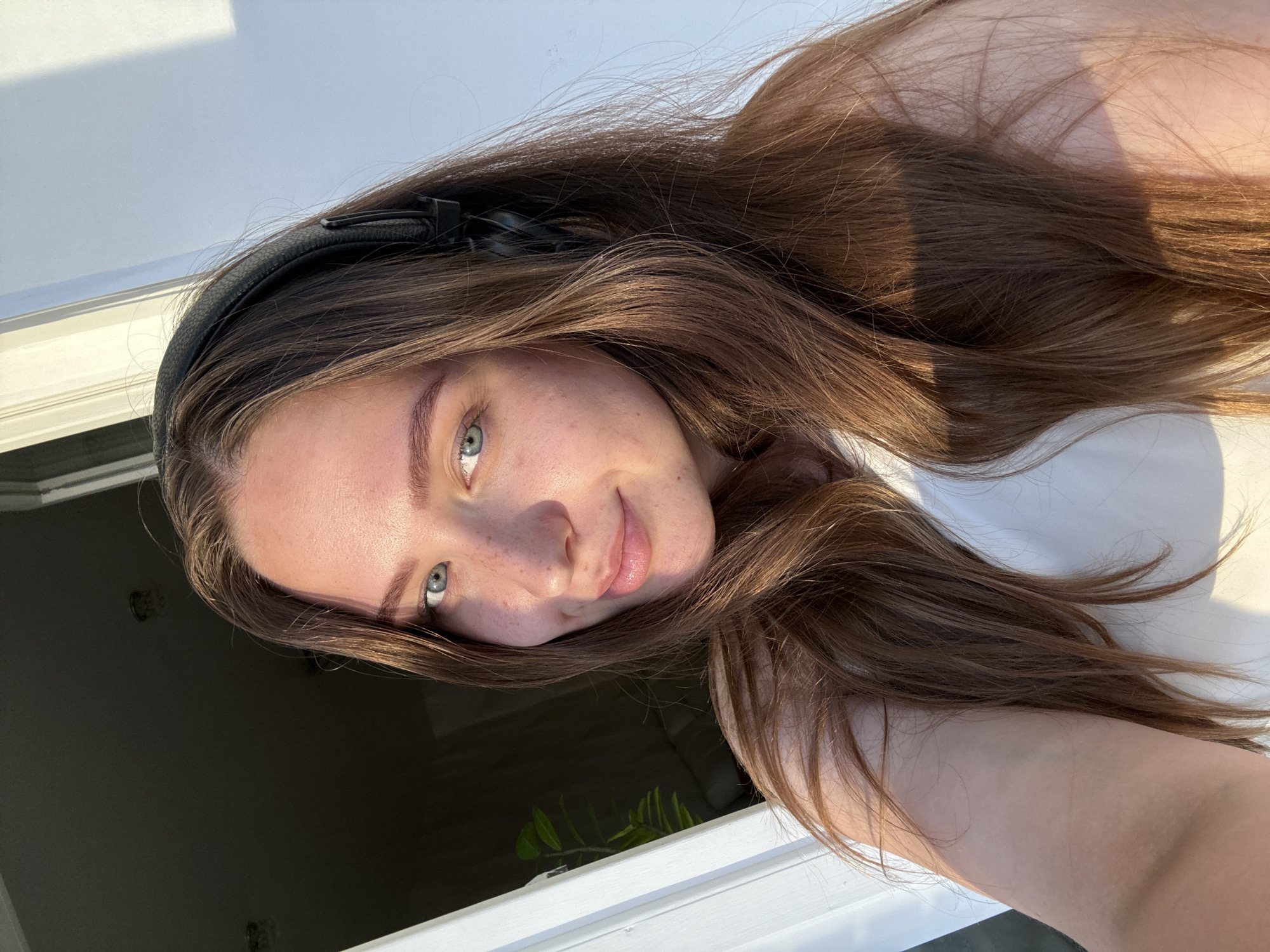 This media file is a selfie of a young woman with long, brown hair. She is wearing black headphones and has a light, happy expression. Her eyes are blue and she has freckles across her nose and cheeks. The lighting suggests it is daytime, likely late afternoon due to the warm, golden hue of the sunlight. She appears to be outdoors or near a window, as evidenced by the bright light and a hint of greenery visible in the background, which could be plants. The woman's arm is visible in the foreground, and she is looking towards the camera with a slight smile. The scene is casual and relaxed, suggesting a personal moment captured by the subject. There is no visible text in the image. The location context of Chaikovsky, Russia, does not provide specific visual cues within the image itself but can be considered as the setting for this photograph.