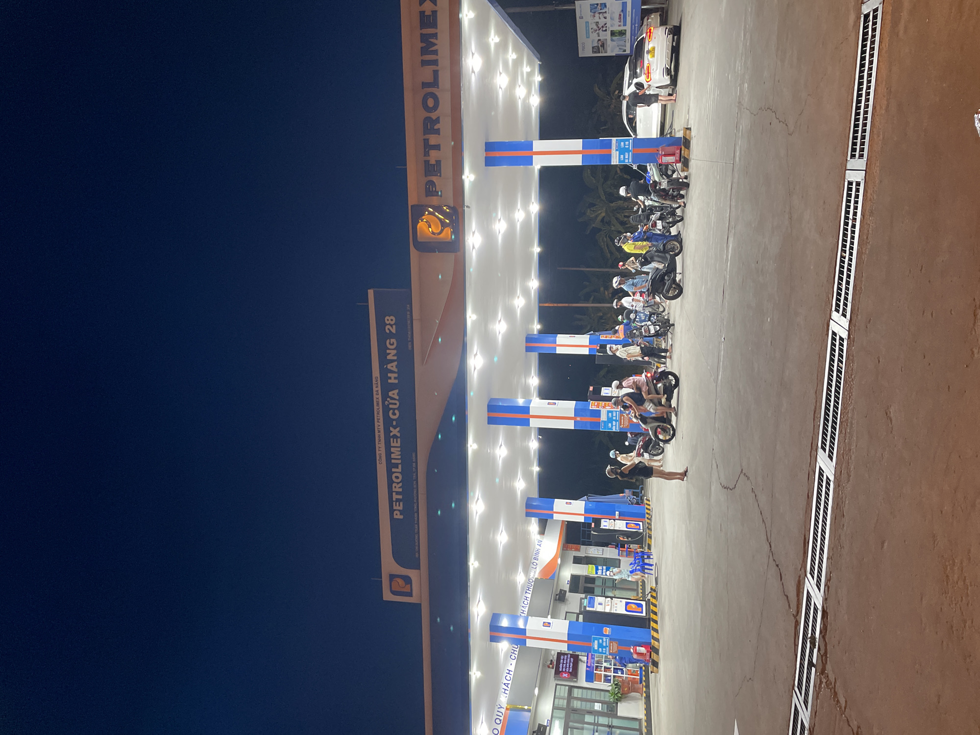 This media file captures a nighttime scene at a Petrolimex gas station in Da Nang, Vietnam. The primary subjects are several individuals on motorcycles, likely refueling or waiting their turn. The gas station itself is brightly illuminated by overhead lights, casting a glow on the pumps and the paved forecourt. A white car is also visible at one of the pumps, with a person attending to it.
The most prominent text visible identifies the location as "PETROLIMEX - CỬA HÀNG 28" (Petrolimex - Store 28) and indicates it is a branch of "CÔNG TY TNHH MTV PETROLIMEX ĐÀ NẴNG" (Petrolimex Da Nang Company Limited). Other text appears to be Vietnamese signage related to customer service or safety, such as "KHÁCH THƯỢNG LỘ BÌNH AN" (Customers have a peaceful journey).
The time of day is clearly night, suggested by the dark sky and the artificial lighting. The weather appears to be clear. The overall atmosphere seems to be one of everyday activity at a busy service station, with a functional rather than emotional tone. The scene is characterized by the brightly lit infrastructure of the gas station, the presence of vehicles, and the people engaged in their activities.