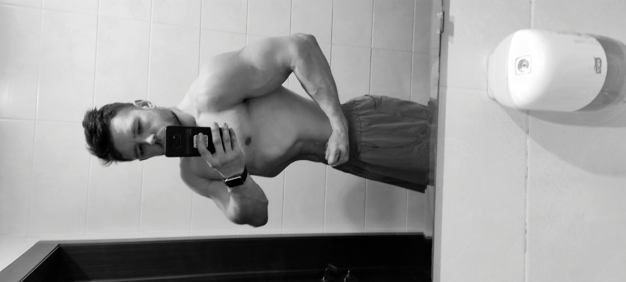 A shirtless man wearing shorts takes a selfie in a bathroom with white tiled walls. The location is presumably Fryazino, Russia. A white Tork dispenser is mounted on the wall. A dark door frame is visible. The man is holding a black smartphone. He is wearing a wristwatch.