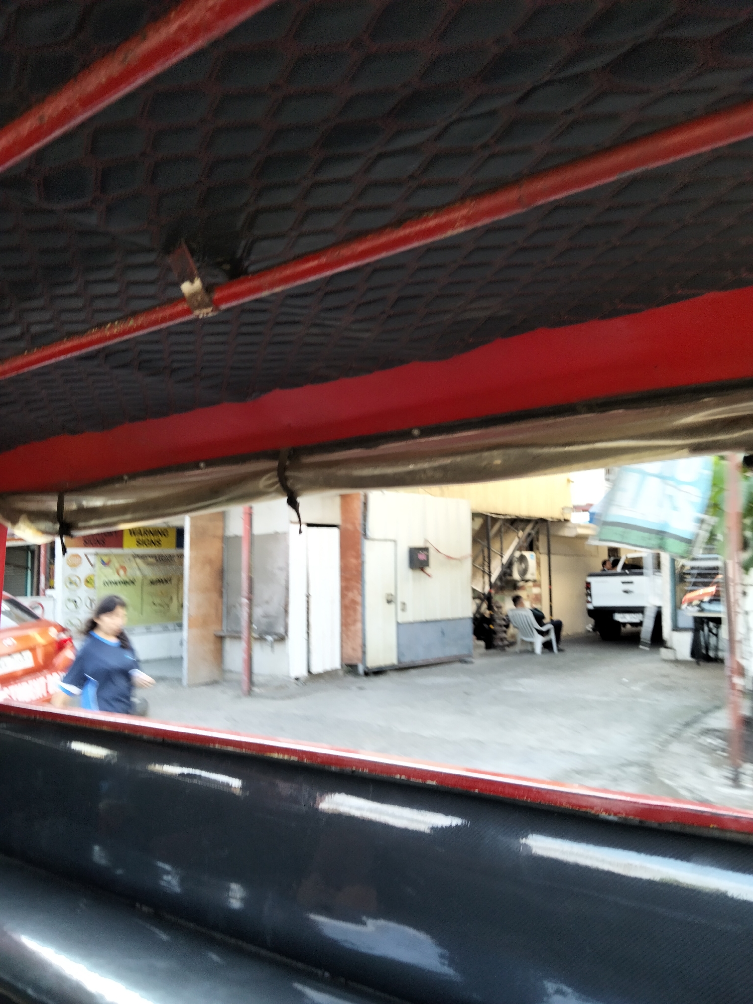 The image captures a street scene in Davao City, Philippines, viewed from a slightly elevated perspective, likely from within a vehicle. The foreground is dominated by a dark, textured surface, possibly the interior of a vehicle's roof or a cargo bed, with red metal framing along the edges.
In the middle ground, a woman in a dark blue shirt with a light blue trim is walking. Behind her, to the left, an orange vehicle is partially visible. Further back, a building with various doors and windows stands. A yellow sign with the words "WARNING SIGNS" is mounted on the wall. Several individuals are seated on chairs outdoors, in front of a building. A white pickup truck is parked to the right of these individuals. The environment appears to be a somewhat industrial or commercial area, with multiple structures and vehicles present. The lighting suggests it is daytime.