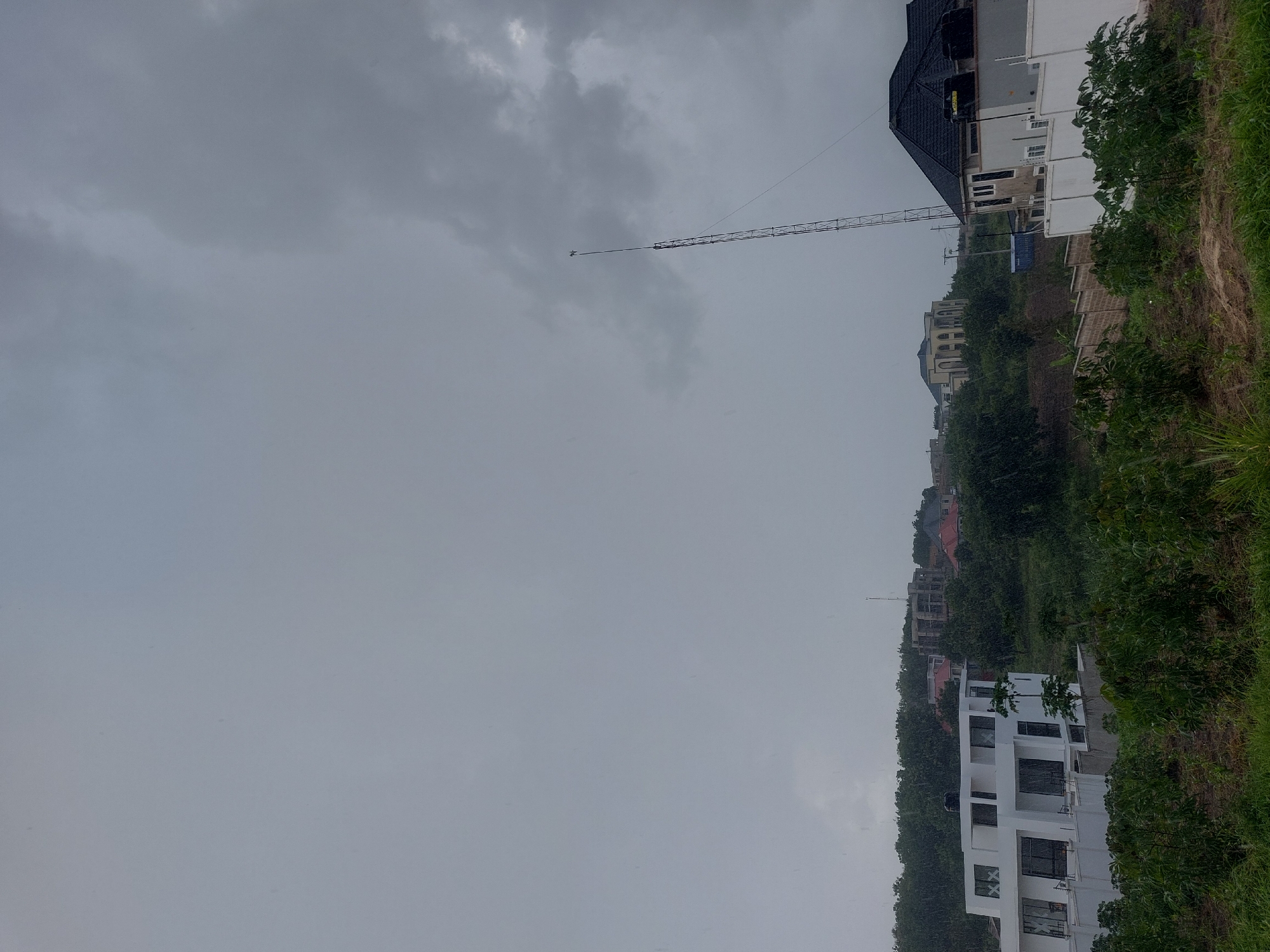 The image depicts a residential area in Dar es-Salaam, Tanzania, under an overcast sky. The scene is characterized by a mix of modern and older style buildings situated on a hillside covered in lush, green vegetation. A communications tower rises into the sky. A few houses are colored in white and cream colors. The weather appears gloomy, with the sky filled with dark clouds and likely rain. The focus is on the urban environment.