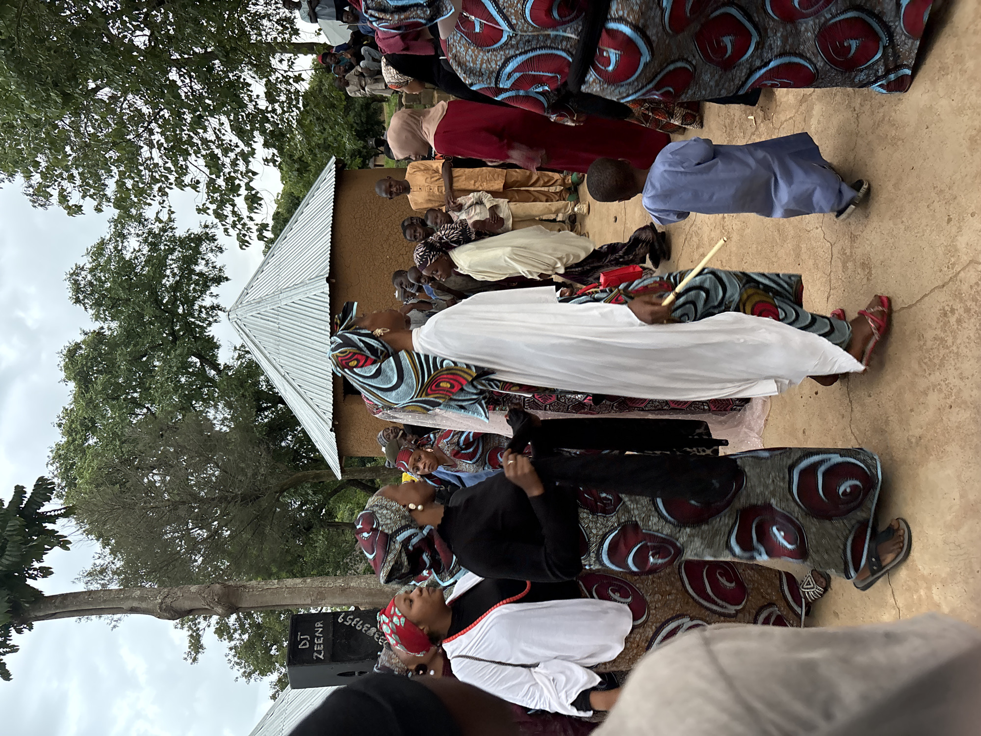 An outdoor gathering is taking place in the city of Jos, Nigeria, with a focus on a group of people dressed in traditional African attire. The central figures are women wearing vibrantly patterned fabric, many with matching headscarves. One woman in the foreground is clad in a flowing white garment over a black top and a patterned skirt. To her left, another woman wears a black top and a similar patterned skirt, adorned with a red beaded necklace. The background reveals a rustic setting with traditional mud-brick huts topped with corrugated metal roofs. A DJ speaker is visible to the left, suggesting music is part of the event. Several children are also present, including a young boy in a blue traditional outfit walking away from the camera. The overall atmosphere appears festive and communal.