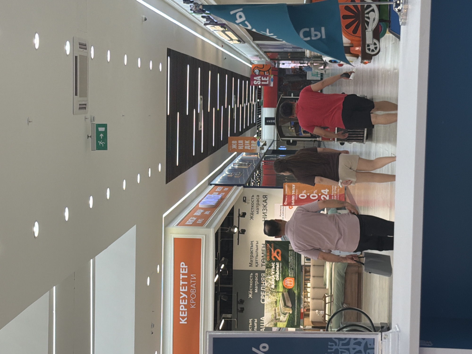 The image depicts the interior of a shopping mall in Astana, Kazakhstan. In the foreground, a person walks away from the camera, carrying a grey bag. Behind them, two other individuals are also walking away from the camera. The mall features bright, modern lighting with linear lights along the ceiling and recessed circular lights. Various storefronts are visible, including a bedding or furniture store with a large advertisement featuring a bed and promotional text. Further down the mall, signs with Cyrillic script are visible, and a colorful toy train can be seen. An exit sign with a green arrow pointing right is also present on the ceiling. The environment appears clean and well-maintained, characteristic of a contemporary shopping complex.