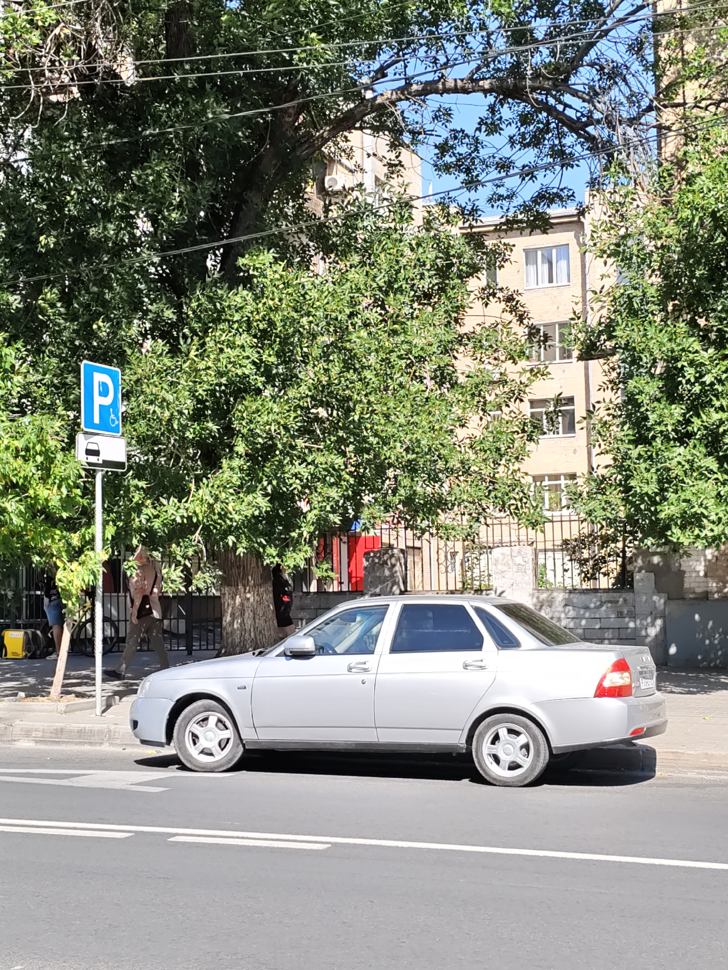 A silver sedan is parked on the side of a street in Rostov-on-Don, Russia. The car is facing towards the right of the frame and its front wheels are turned slightly. A pedestrian crossing is visible in the foreground. To the left of the car, a blue and white parking sign with a wheelchair symbol and a car icon is mounted on a pole. Behind the car and sign, there is a large tree with dense green foliage. In the background, a light-colored building with multiple windows can be seen, partially obscured by the tree. Two people are walking on the sidewalk to the left of the parking sign. The scene appears to be on a sunny day.