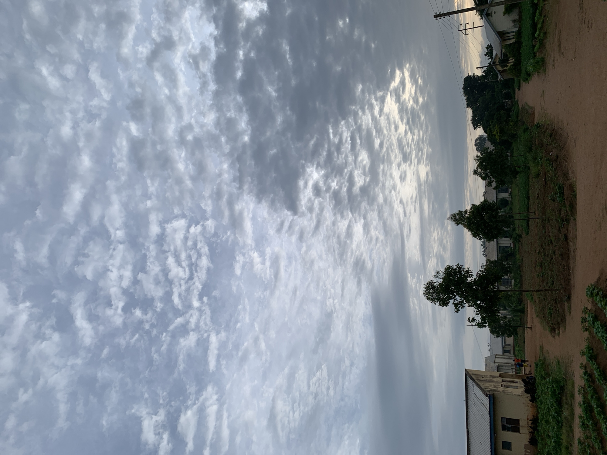 This is a vertical, eye-level, medium shot photograph taken in Kuru Karama, Nigeria. The image captures a cloudy sky with scattered white and gray clouds, extending across the entire upper half of the frame. In the foreground and midground, there are several buildings with light-colored walls and metal roofs. Trees and vegetation are visible around the buildings, and a dirt path runs through the scene. Two people, appearing to be children or young adults, are walking on the path near the left side of the image. In the far right of the image, there are utility poles with wires. The overall lighting suggests it may be early morning or late afternoon.