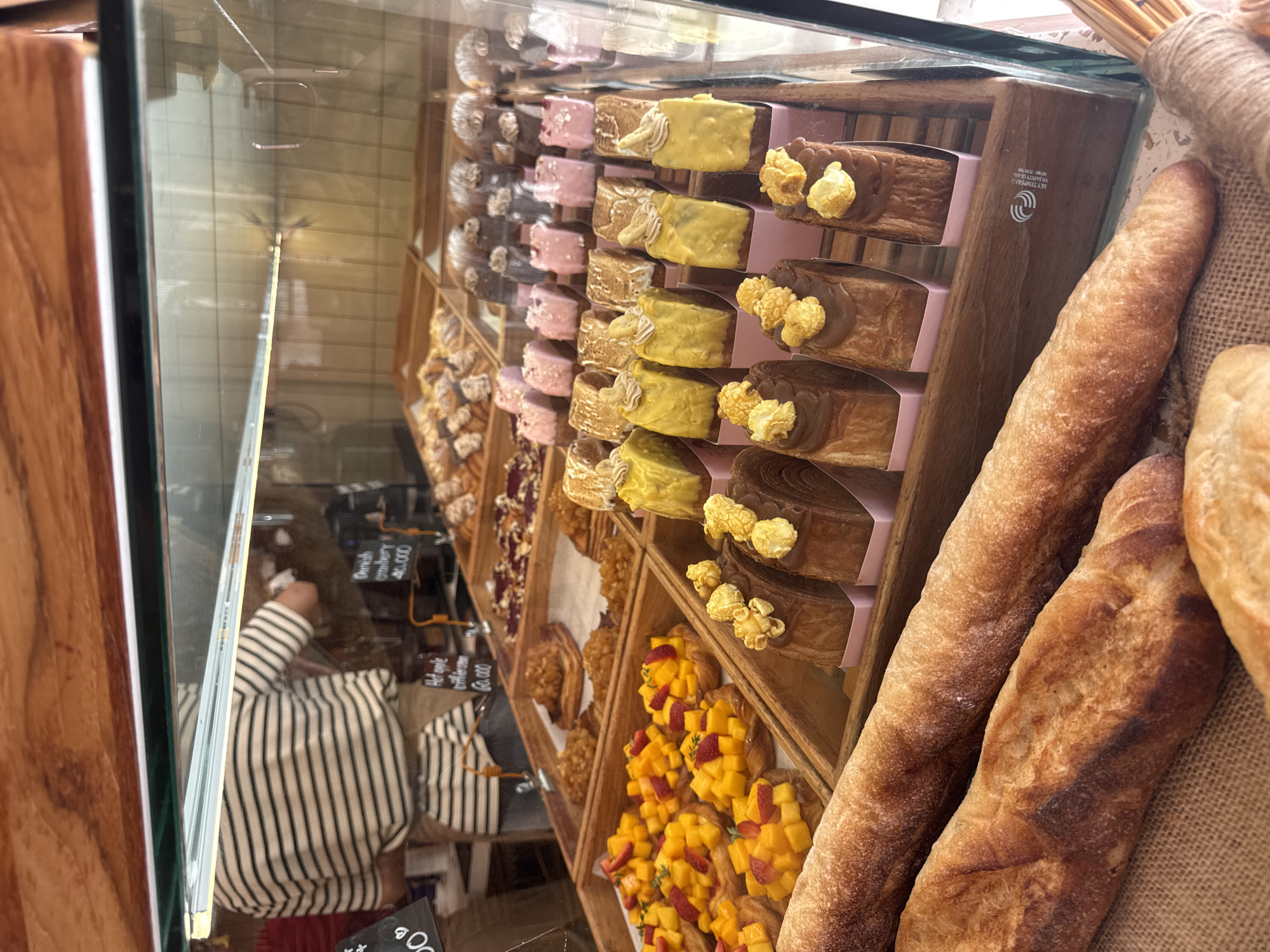 The image shows a close-up view of a bakery display case in Nha Trang Ward, Vietnam. The display case is filled with various baked goods, including baguettes, pastries with mango and strawberry toppings, chocolate-covered pastries topped with popcorn, and smaller pastel-colored pastries. In the background, a person wearing a striped shirt is partially visible, appearing to be a baker or staff member. The display case is made of glass and wood, and the lighting suggests an indoor setting. The overall scene depicts a commercial establishment offering a selection of sweet and savory baked items.