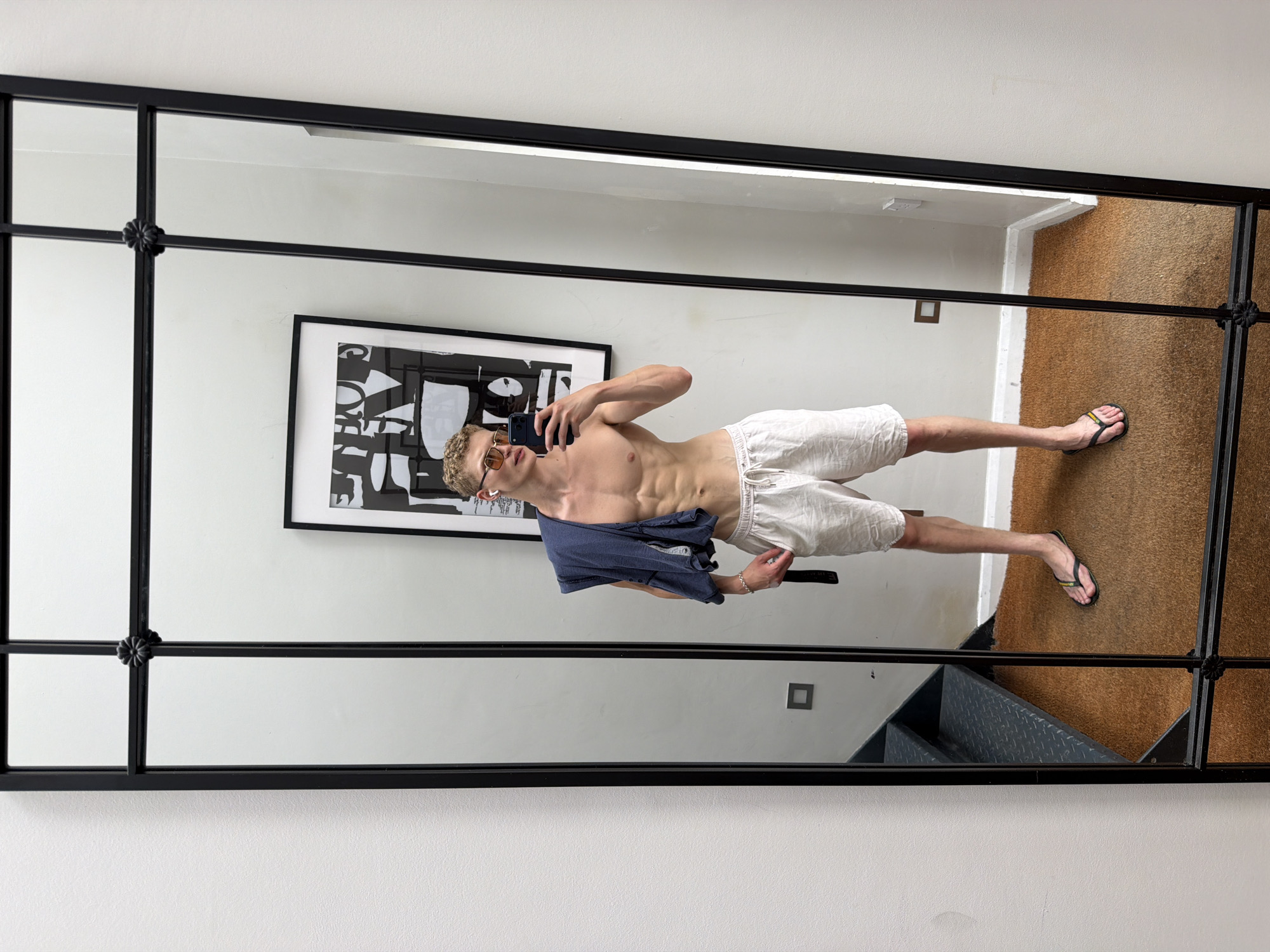 A young man with blonde hair and a muscular physique is taking a selfie in a large, multi-paneled mirror. He is wearing light-colored linen shorts and flip-flops, and has a dark blue t-shirt draped over his left shoulder. He is also wearing sunglasses. The mirror reflects a room with a light-colored wall, a framed black and white abstract print, a dark-colored staircase with metal treads, and a brown rug. The man appears to be standing on the rug in what looks like a dressing room or a hallway. There are electrical outlets visible on the wall. The image does not provide any information about the location being in London, United Kingdom.