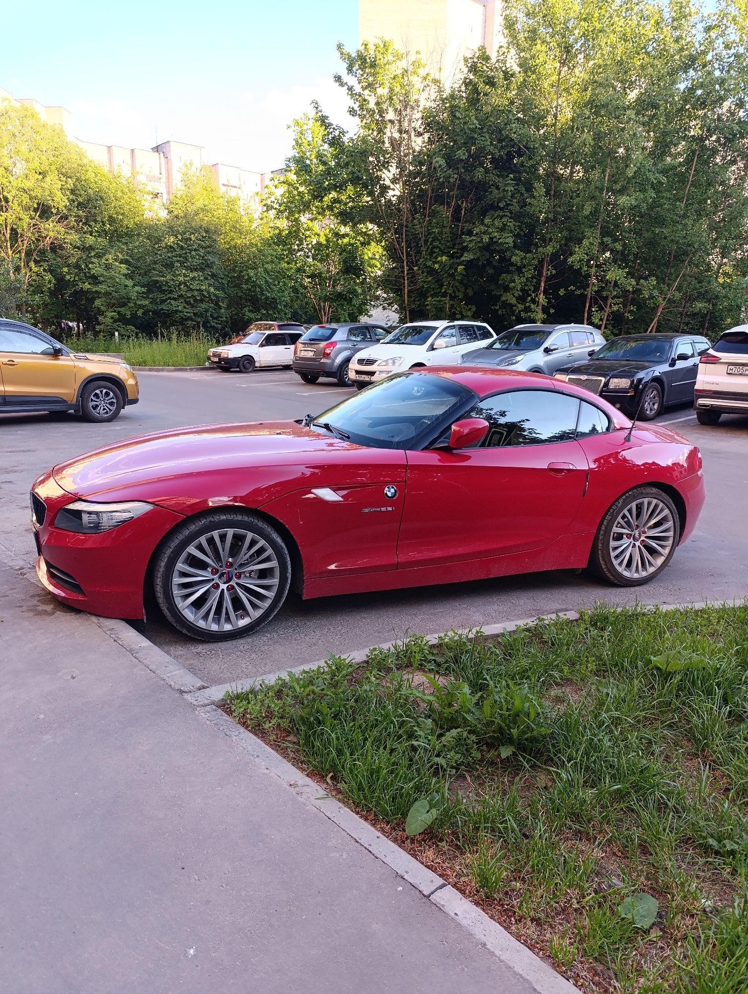 A vibrant red BMW Z4 E89 hardtop convertible sports car is the prominent subject, parked on the edge of a paved lot. Its front left wheel is slightly over a concrete curb separating the parking area from a strip of unkempt grass. The car features distinctive multi-spoke alloy wheels with red accents on the hubcaps and bears an "sDrive35i" badge on its side fender.
The scene depicts an outdoor parking area during a bright day, likely late afternoon or early morning, given the soft, natural sunlight and clear, light blue sky. In the background, numerous trees with dense, green foliage partially obscure multi-story residential buildings.
Several other vehicles are parked behind and to the left of the red BMW. These include a partial view of a gold/yellow SUV on the left, followed by a white sedan, a silver SUV, a black sedan (resembling a Chrysler 300C), and a white SUV further in the background. Visible license plates on some of these vehicles display Cyrillic characters, such as "M705P" on the white SUV in the far background, indicating the location is likely in Russia or a neighboring country. No people are visible, and the setting appears calm and still.