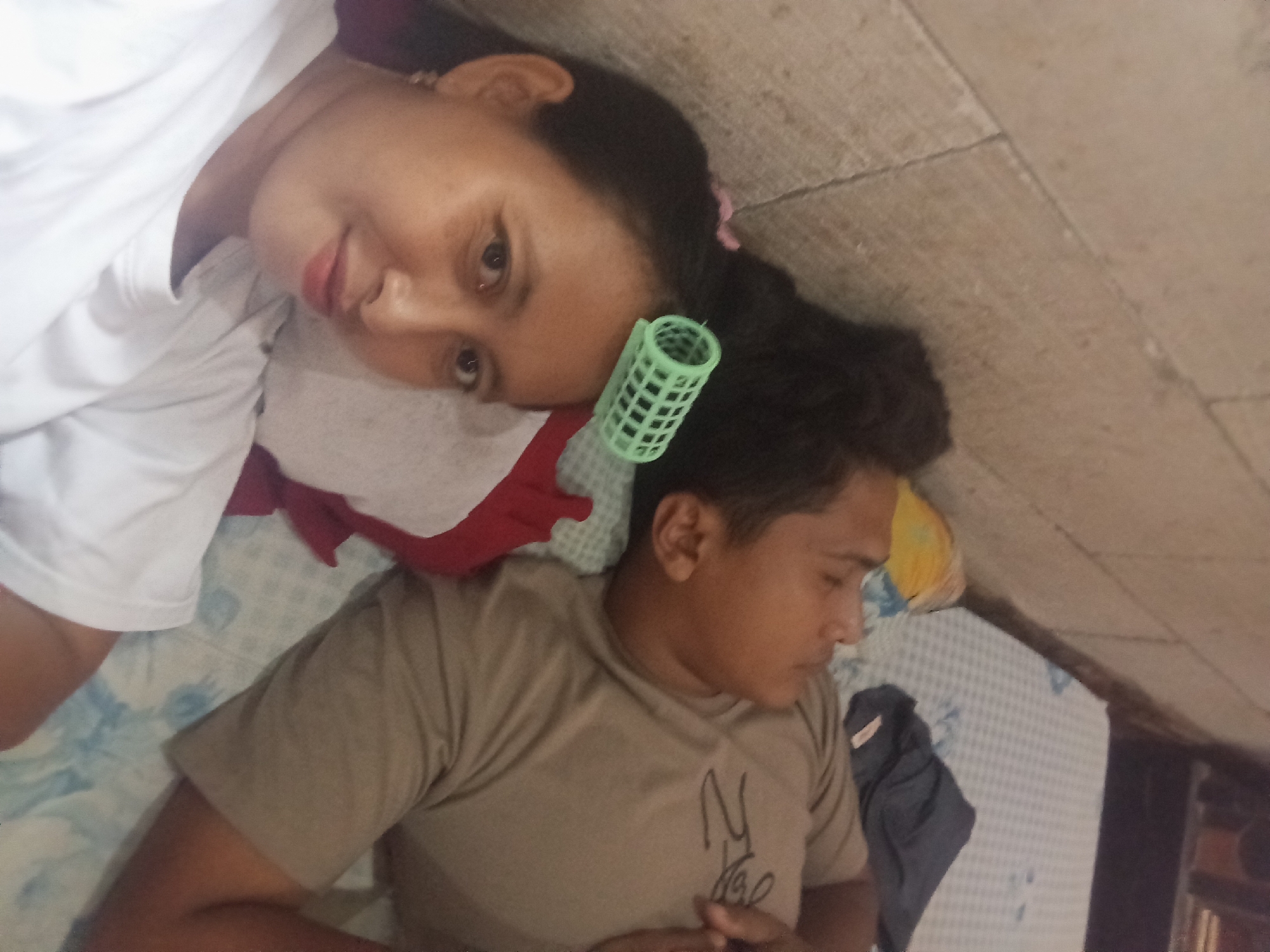 The image depicts two individuals lying on a bed in what appears to be an indoor setting. The individual on the left is a woman with dark hair, and she is wearing a white shirt. A green hair roller is visible in her hair. Her gaze is directed towards the camera. The individual on the right appears to be a man wearing a tan shirt with writing on it. He is sleeping, with his hands clasped near his chest. The bed is covered with a patterned blue and white sheet. The background shows a wall and a partial view of what looks like a cabinet. The image was taken at an angle, with the camera positioned above the subjects. While I am unable to verify the exact location in Baron, Indonesia based on the image alone, the indoor setting and the presence of the individuals are evident.
