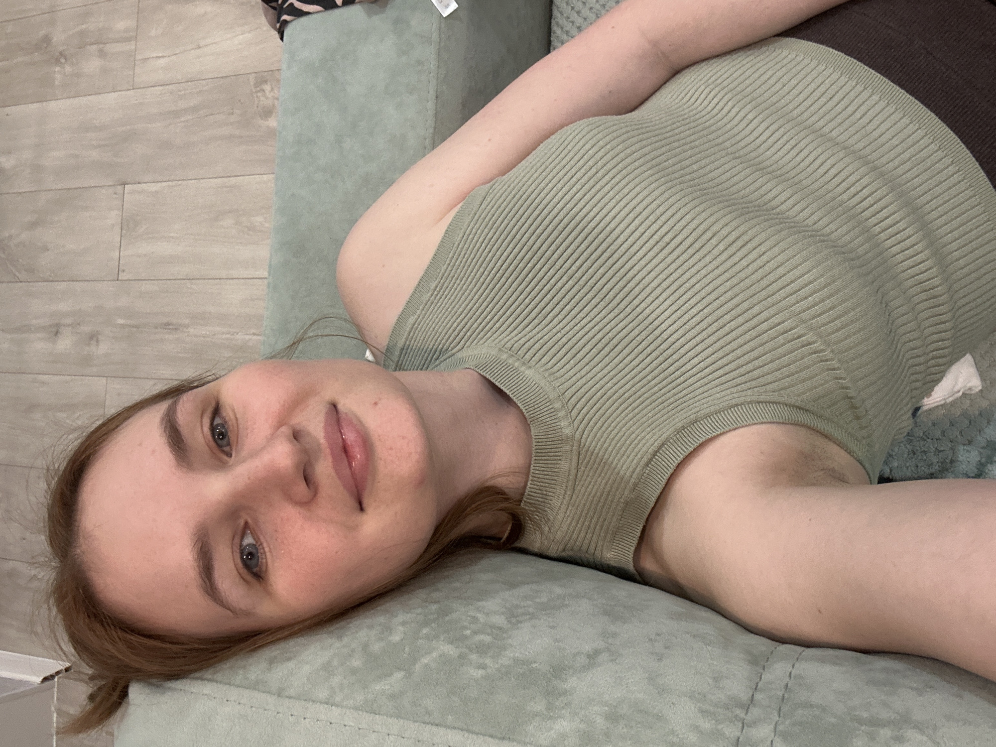 A young woman is depicted lying on a light green sofa indoors, looking towards the camera with a gentle, relaxed smile. She appears to be in her 20s or early 30s, possessing fair skin, light brown or auburn hair, and clear blue eyes. She is dressed in a light sage-green, ribbed sleeveless top, with a dark brown or black garment partially visible behind it.
The setting is an indoor space, likely a home in Chaikovsky, Russia. To the left of the sofa, a light-colored wooden floor with distinct planks is visible. The sofa itself is a soft, cushioned piece of furniture in a muted green hue. In the upper right corner of the frame, a fragment of patterned fabric, possibly black and white, along with a white tag containing unreadable text, can be seen. A small white box or container is also partially visible in the bottom left. The lighting is soft and even, contributing to the casual and comfortable atmosphere of the scene. The woman's relaxed posture and pleasant expression suggest she is at ease, possibly taking a selfie or being photographed while resting.