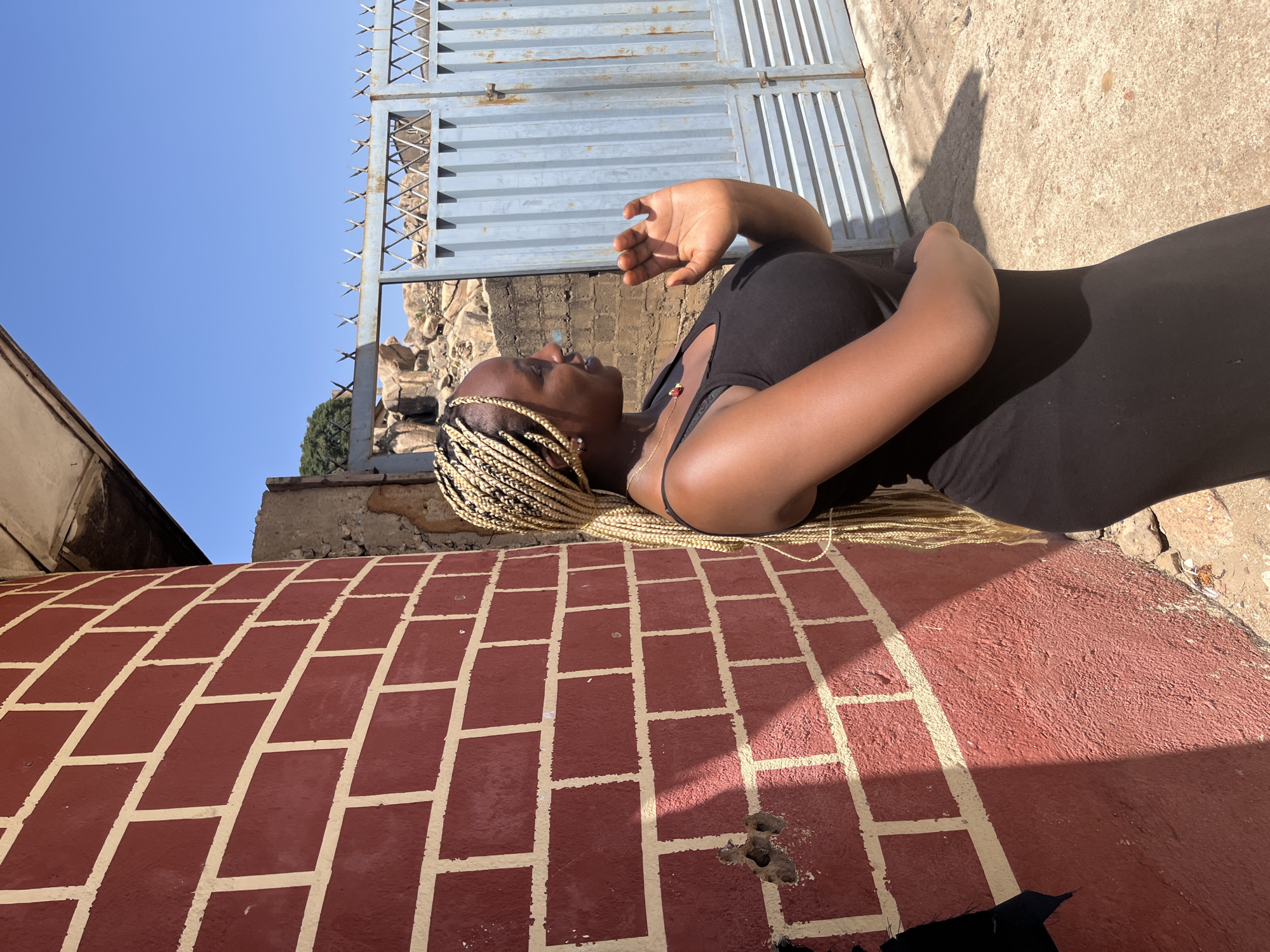 A woman with blonde braided hair is lying on a brick surface under the bright sunlight in an outdoor setting. She is wearing a black tank top and dark pants or a skirt. Her arm is raised in the air. Behind her is a metal gate with spikes and a partial view of buildings or rock formations. The brick surface has a red color with cream-colored grout lines painted on it. Some debris is scattered near the bottom edge of the frame. The sky is visible above the gate and is a clear blue color.