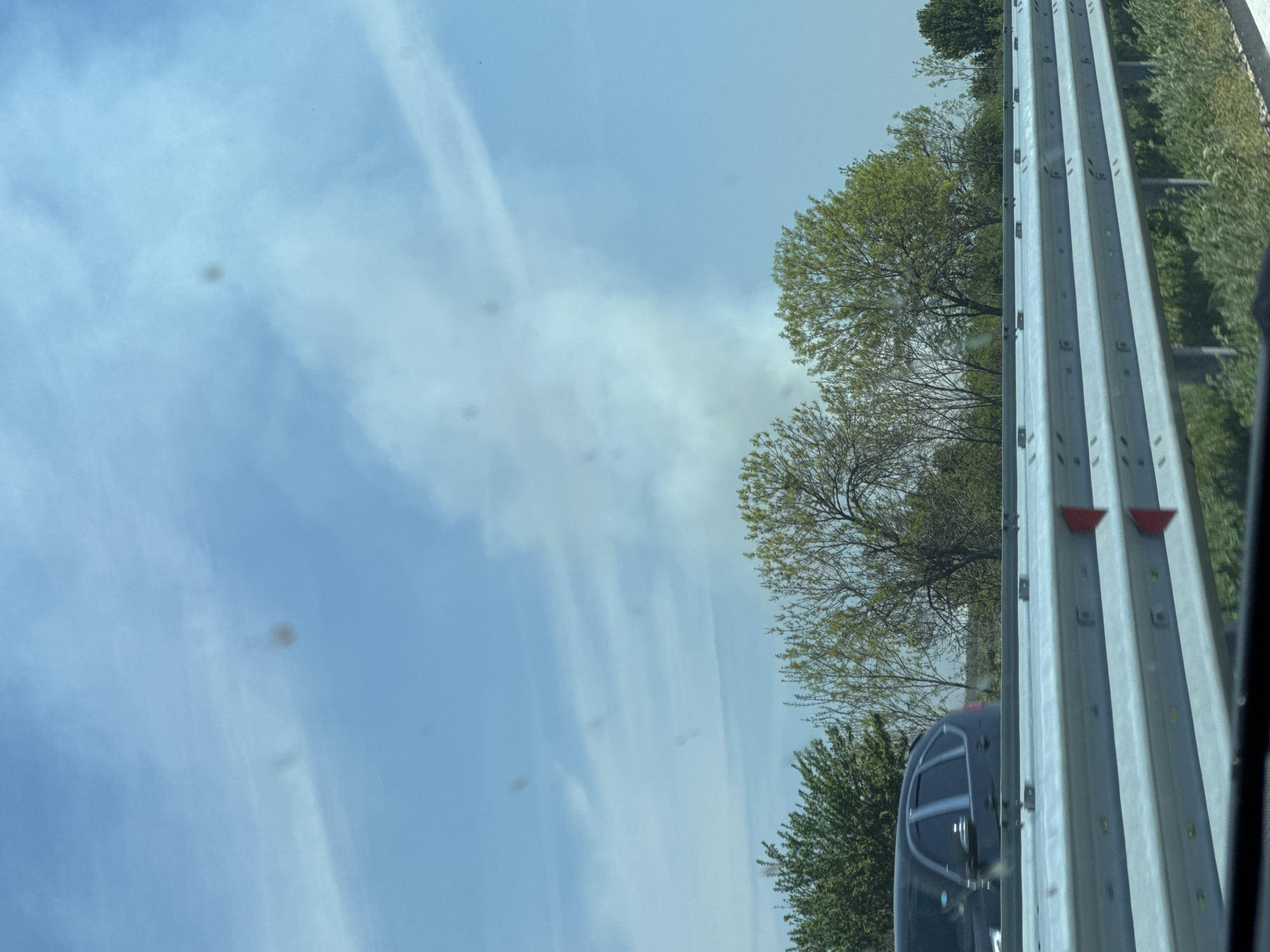The image displays a bright, daytime scene, likely captured from inside a moving vehicle looking out towards the roadside. The sky occupies a large portion of the frame, showing a clear, light blue expanse with several thin, wispy white clouds or contrails stretching across it.
On the right side, a sturdy metal guardrail, composed of several horizontal beams and vertical posts, runs from the foreground into the distance. It is light grey or silver in color, and two red, triangular reflectors are visible on its uppermost rail. Beyond the guardrail, verdant grass and trees with full green leaves line the roadside, suggesting a spring or summer season. Another tree in the mid-ground appears to have sparser foliage.
A partial view of a dark-colored vehicle, possibly a car or SUV, is visible in the lower right, positioned behind the guardrail.
A notable detail throughout the sky are numerous dark, blurry, and out-of-focus spots of various sizes, appearing as smudges or dust on the camera lens or the window through which the photograph was taken.
The lighting indicates it is daylight, under a clear or partly cloudy sky. There are no people visible, nor any discernible text. The scene depicts a moment of travel, typical of a highway or major road environment. While the location context of Malo Konare, Bulgaria, is provided, there are no specific visual cues within the image to confirm this.