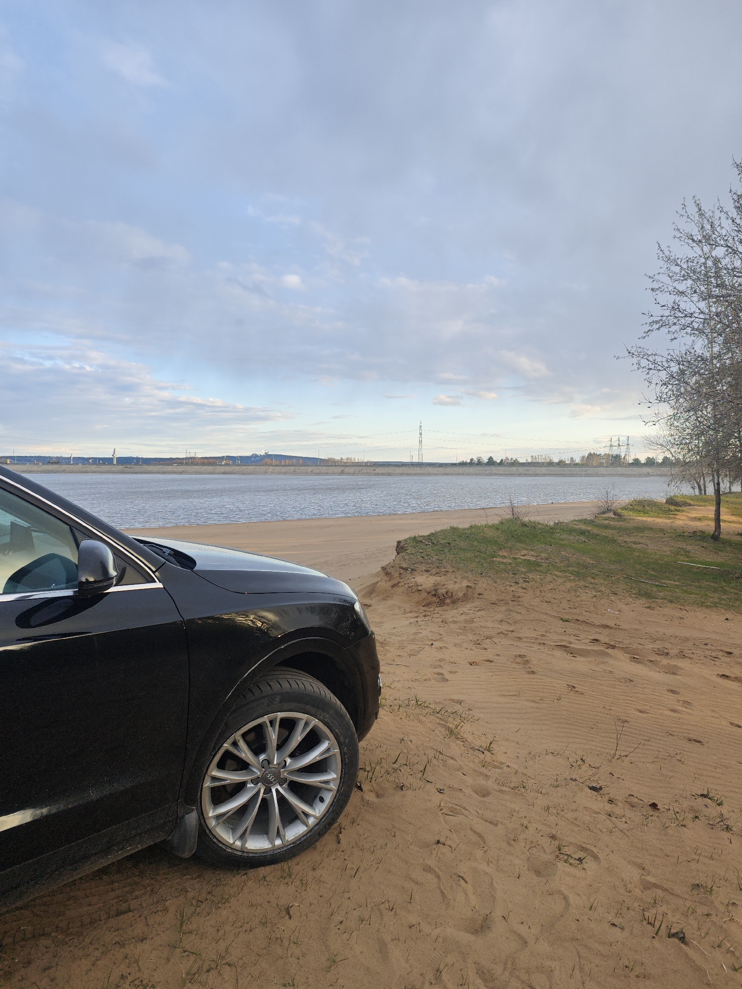 A black Audi SUV is prominently featured on the left side of the frame, parked on a sandy beach. Only the front left quarter of the vehicle, including its wheel with the visible Audi logo, is visible. The sandy ground extends to the right and foreground, marked with footprints and sparse patches of green grass. A small, grassy dune separates the immediate sandy area from the water's edge.
Beyond the sand and grass, a wide body of calm water stretches across the mid-ground, likely a river or large lake, given the location context of Noviy, Russia. The far bank across the water shows a low-lying horizon with various distant structures, including power lines and several tall transmission towers, suggesting industrial or infrastructure presence. Bare trees or trees with minimal foliage stand on the far right of the sandy area, adding to the natural elements.
The sky is overcast with soft, diffused light, indicating a cloudy day, possibly late morning or afternoon. The overall atmosphere is tranquil. The weather appears mild with no signs of rain. The scene combines natural elements like sand, water, and trees with the man-made car and distant industrial infrastructure.