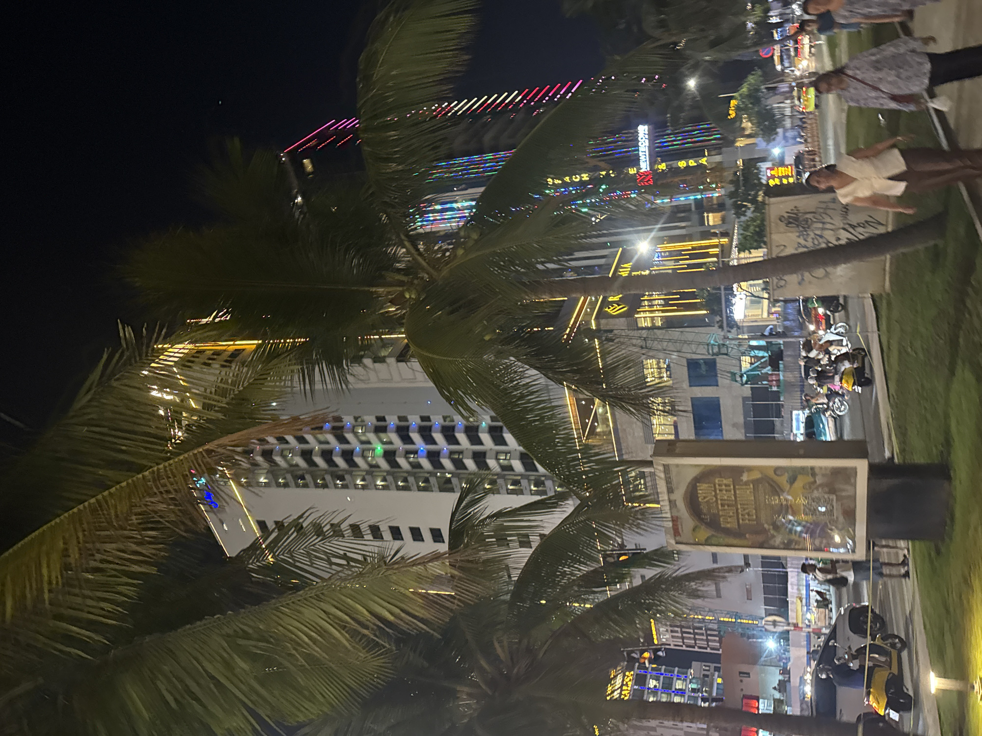 The image is a night scene in Đà Nẵng, Vietnam. Palm trees are in the foreground. In the background are city lights, including lights atop and on buildings. People are walking along the sidewalk, and motorbikes are parked and driving nearby. A sign advertising the "Sun Kraft Beer Festival" is visible.