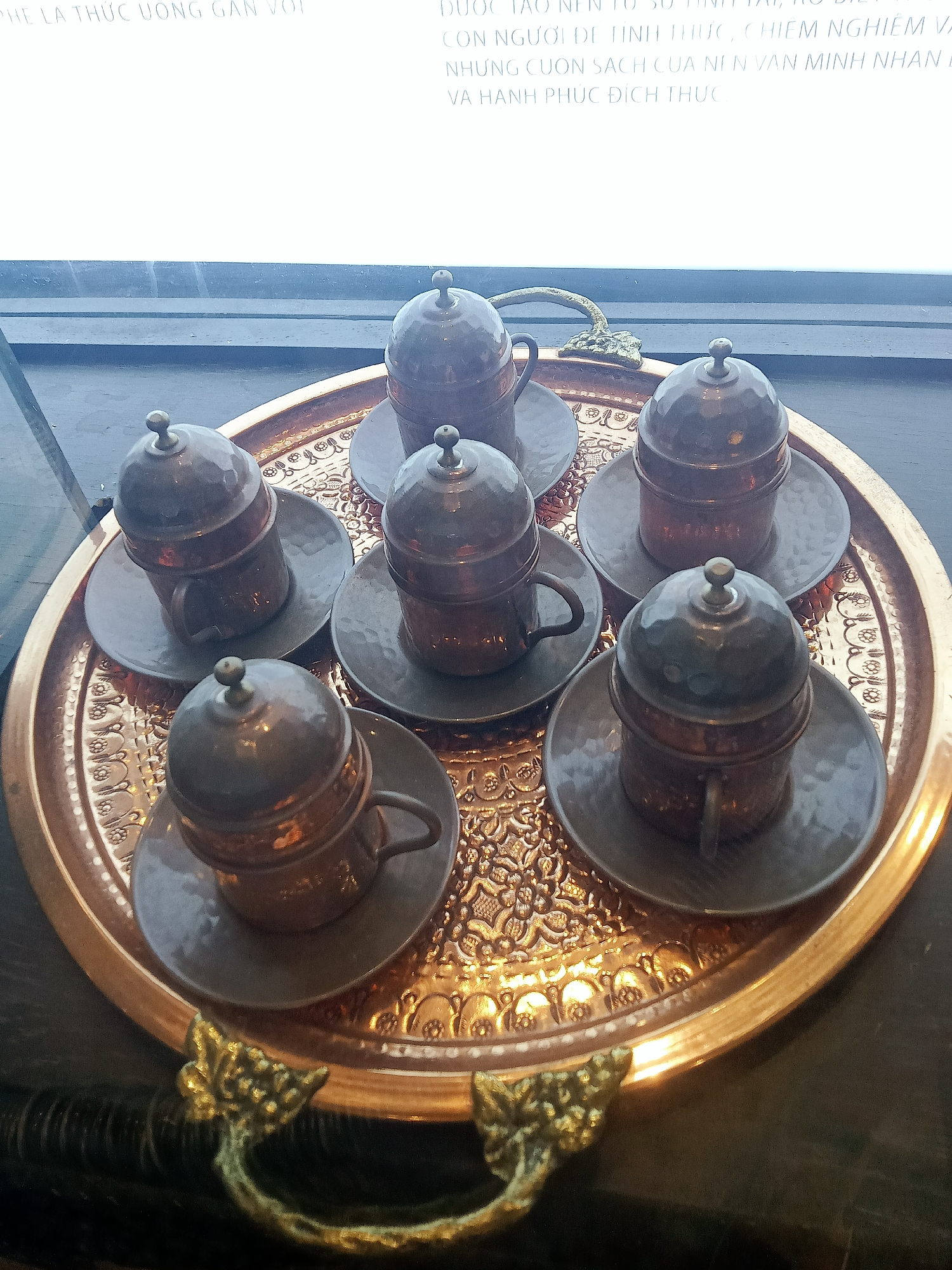 The image shows a copper tray with six small, covered cups and saucers on a dark surface. The tray has ornate handles and a detailed, textured surface. The cups are arranged in a circular pattern around the center of the tray. There is text in Vietnamese visible at the top of the image, likely part of a sign or display. The scene appears to be indoors, near a window or light source. Based on the metadata information available (though not directly visible in the cropped images), this scene is likely located in Nha Trang Ward, Vietnam.