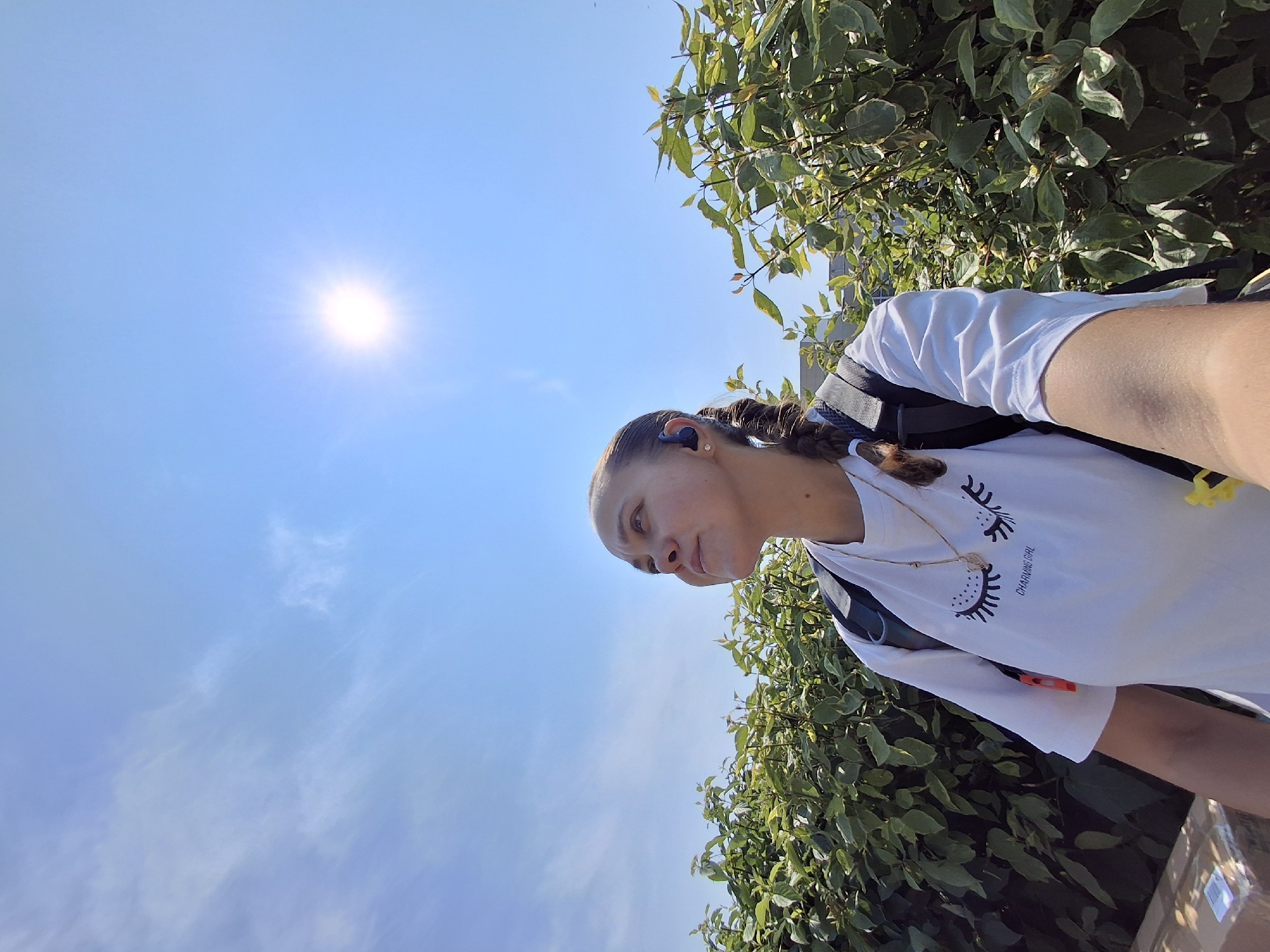 A young woman stands outdoors in front of a dense green hedge, looking upwards. The sky is a bright, clear blue with the sun prominently visible and radiating light. The woman has her hair in a braid and is wearing a white t-shirt with a design of closed eyes and eyelashes printed on it, along with the text "Charming Girl". She also wears a gold necklace and appears to have a black backpack on. The scene is captured from a low angle, suggesting the photo was taken by the woman herself, possibly using a selfie-style shot. The presence of a cardboard box in the foreground suggests an urban or suburban environment. The location is not explicitly mentioned in the image or its crops.