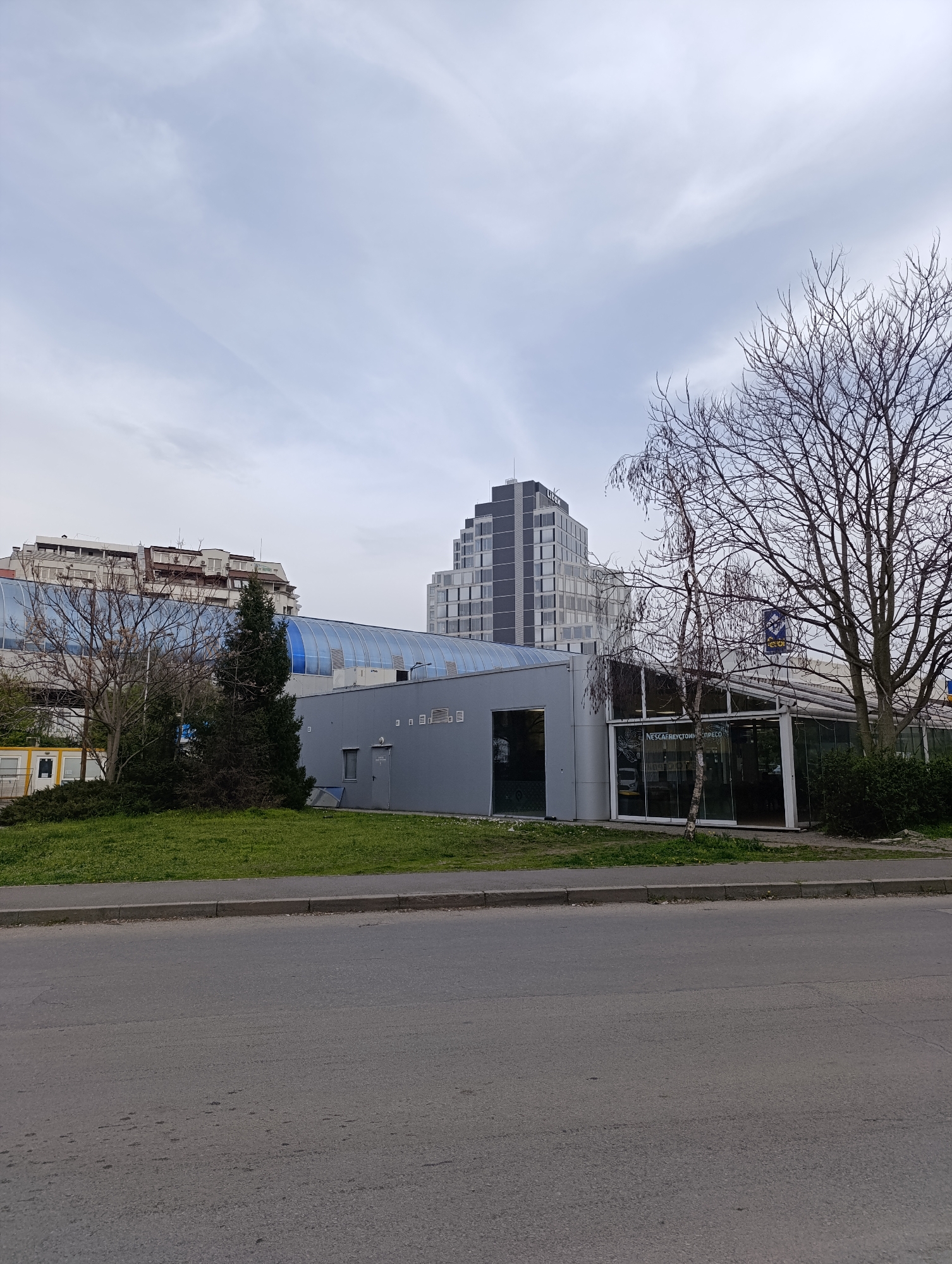 The image shows a street scene in Sofia, Bulgaria. In the foreground is an asphalt road with a curb. Beyond the curb is a grassy strip with some bushes and small trees. Behind that is a gray, modern-looking building with large glass windows and doors. A blue, translucent walkway connects the gray building to a taller building with what appears to be residential units. In the background is a modern, stepped high-rise building. The sky is overcast. A tree with bare branches is on the right of the frame. Based on the visible signage and architectural style, this location appears to be near a Nescafé To Go outlet, and the taller building in the background is likely a residential or commercial structure in the city.