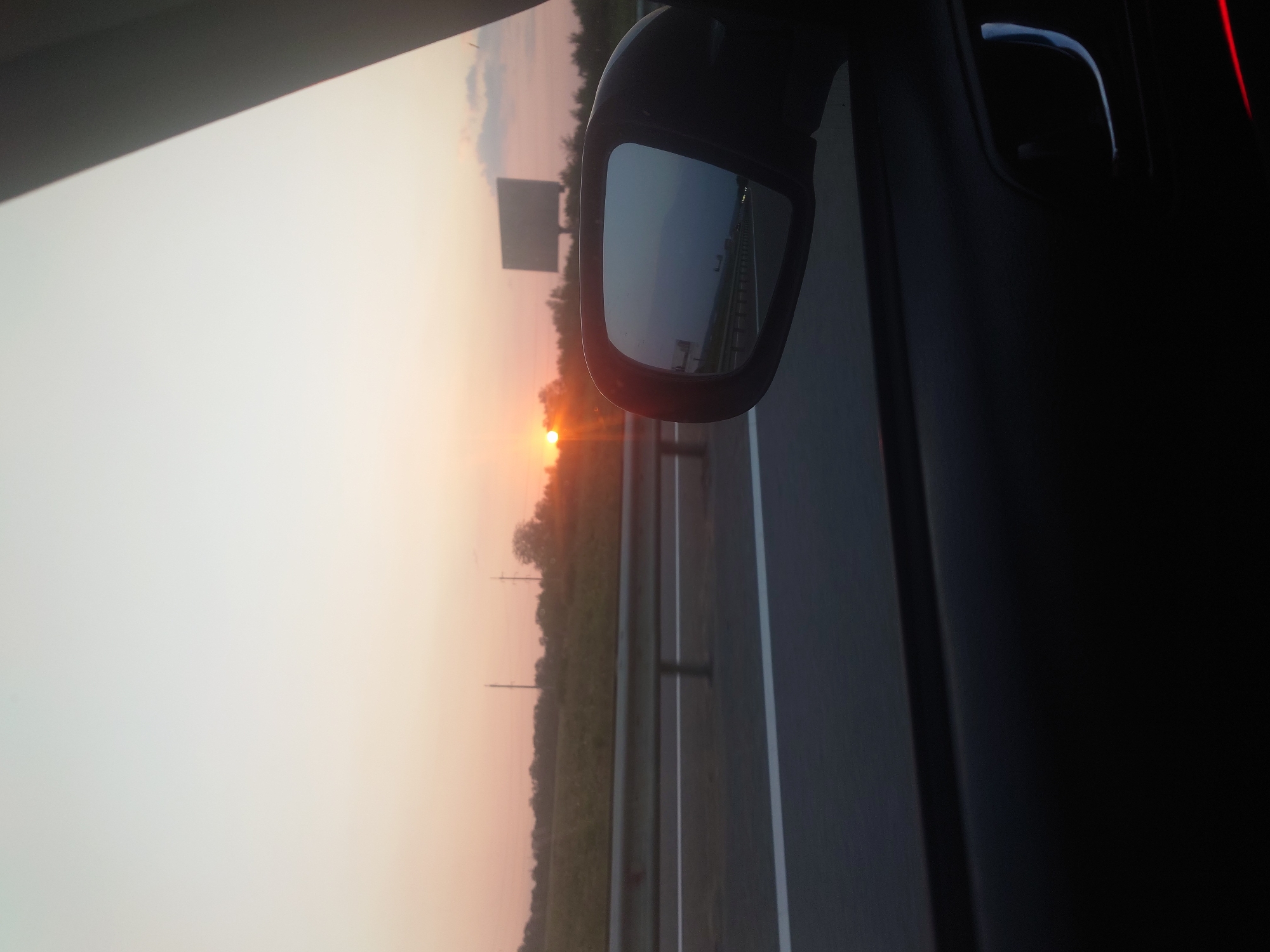 The image captures a sunset view from inside a vehicle, likely a car, driving on a highway. The perspective is from the passenger side, looking out the window and also at the side-view mirror. The sky is a gradient of pale yellow and orange, indicating the sun is low on the horizon, casting a warm glow. In the distance, silhouetted trees form a line along a field, and power lines are visible. A large billboard stands in the background on the right side of the frame.
The side-view mirror reflects the scene behind the car, showing the highway, guardrails, and the receding landscape under the sunset sky. The interior of the car is visible in the foreground, with the dark door panel and the door handle. The road surface is dark asphalt with white lane markings. The overall impression is one of travel during the golden hour.
The location is described as Sokolohirsk, Ukraine. Given the highway and the rural landscape, it is likely a view from a road approaching or leaving the city. The time of day is sunset. There are no people or specific interactions depicted. The objects present are the car's interior, the side-view mirror, the highway, a guardrail, fields, trees, power lines, and a billboard. The environment is a highway setting during sunset.