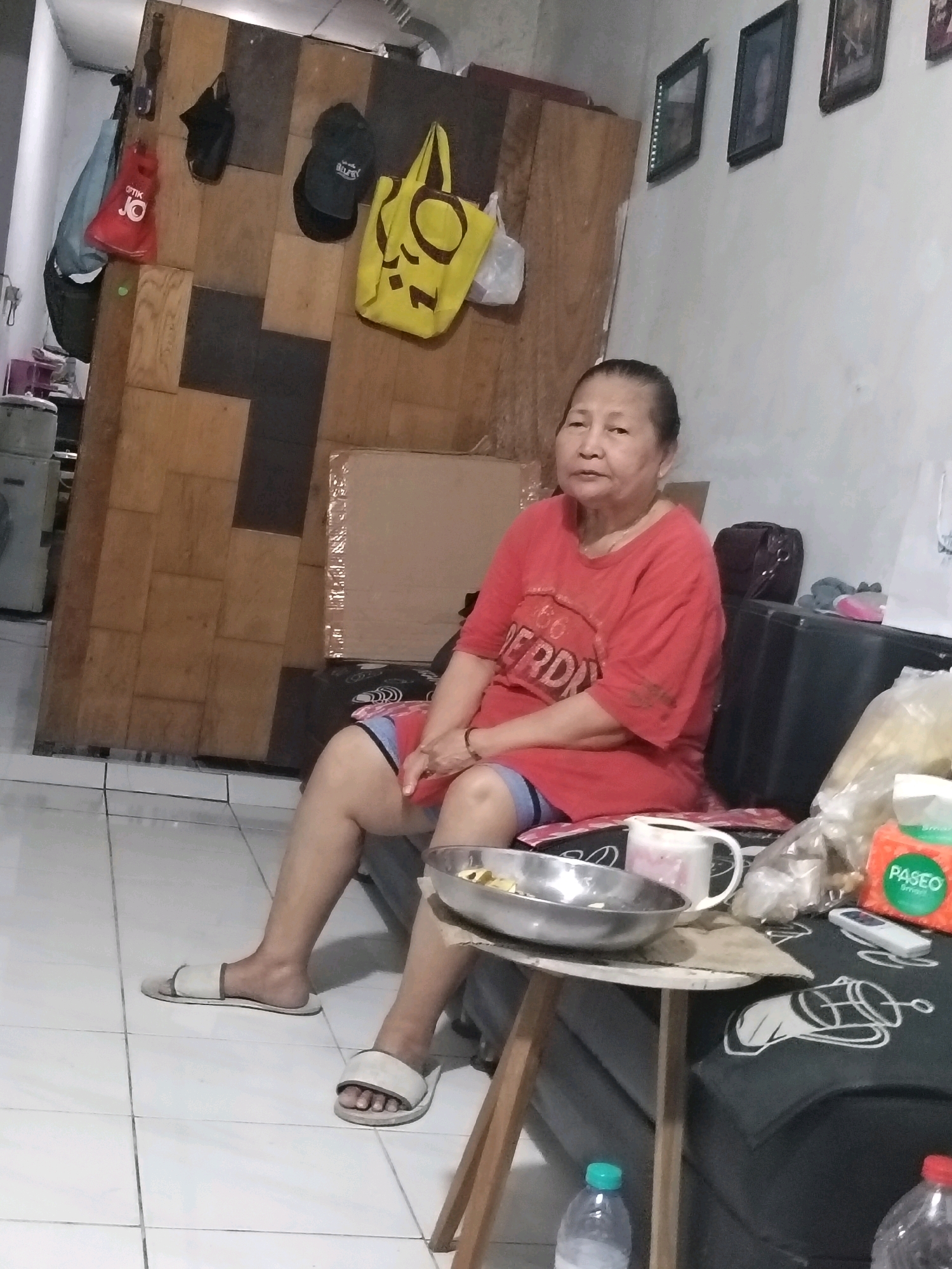 An elderly Asian woman sits on a couch in a dimly lit room. She is wearing a red t-shirt and blue and red checkered shorts. She has on white slippers. In front of her, on a small wooden table, is a metal bowl, a white pitcher with a pink liquid, and a package of tissues. The woman is looking at the camera with a neutral expression. The room appears to be a living room with a wooden partition on the left side of the image, and framed pictures on the wall behind the woman. There is also a large black storage container to the right of the woman. The floor is tiled. The room is cluttered with various items, suggesting it is a lived-in space. The overall atmosphere is informal and domestic.