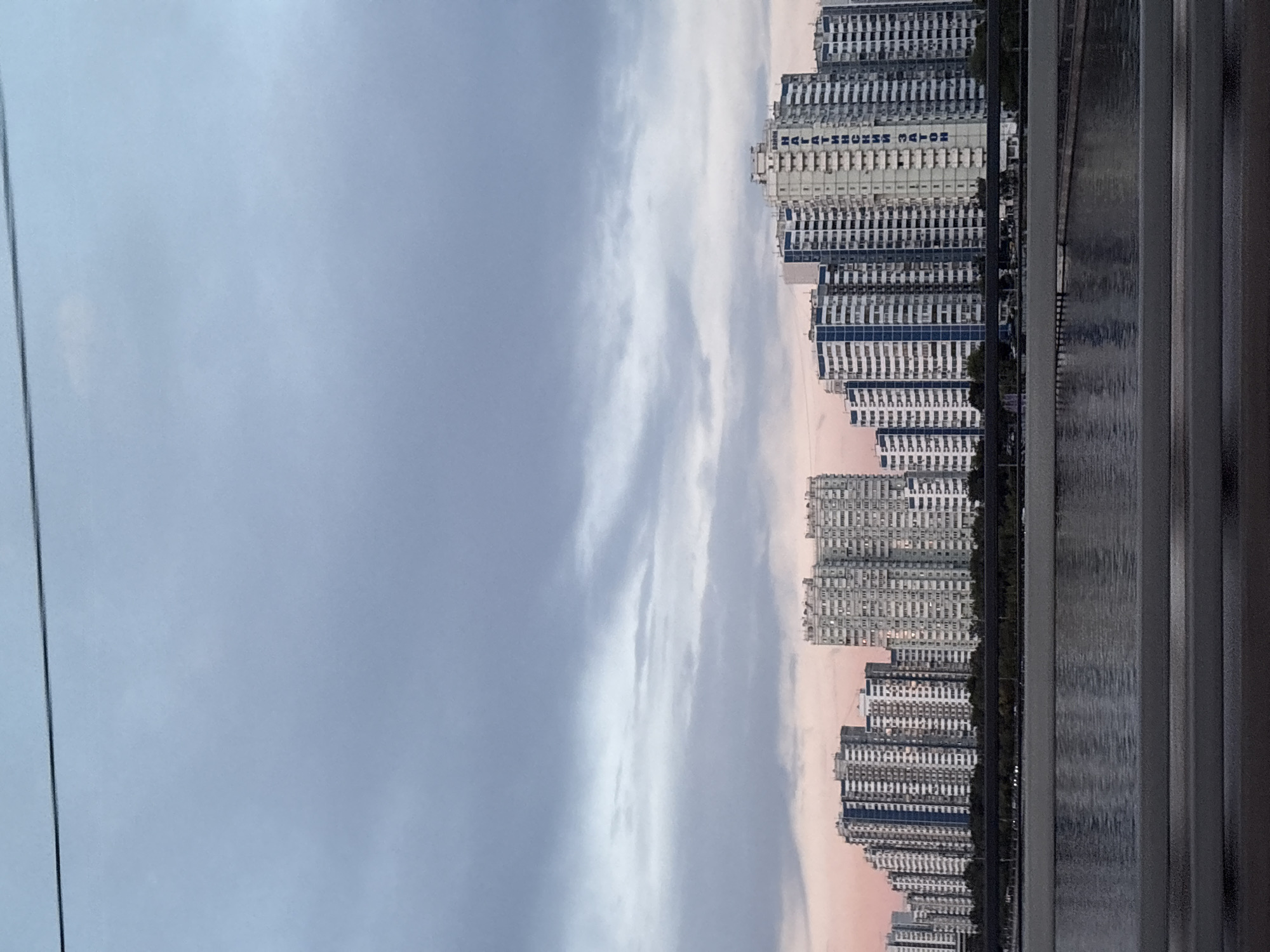 The image shows a row of tall, modern apartment buildings lined up along a body of water. The buildings are white with dark blue balconies and windows, creating a repetitive architectural pattern. The sky above is cloudy, with streaks of pink and orange suggesting it may be sunset or sunrise. There is a reflection of the buildings on the water's surface. A dark, vertical line, possibly a cable or wire, runs down the left side of the image. In the foreground, on the right, there are dark, vertical lines that appear to be part of a window frame or balcony railing. Some text is visible on one of the buildings, appearing to be "MALEMUCHO MATOS". The overall scene conveys a sense of urban living by the water.
