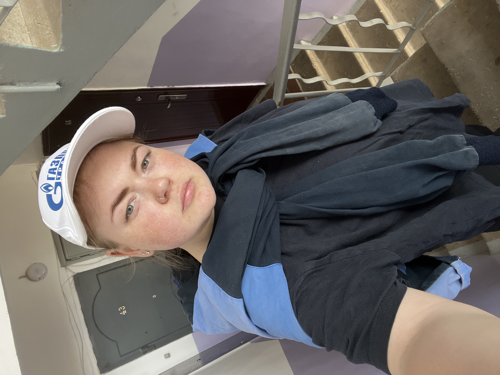 A young woman stands in a stairwell, wearing a white baseball cap with the Gazprom Neft logo, a black shirt with blue sleeves, and a dark blue scarf draped around her neck. She has a nose ring, multiple earrings, and fair skin with freckles. She is holding the camera, taking a selfie. Behind her, the door to apartment number 43 is visible, along with a light fixture on the wall. The stairwell has purple and white walls, with metal railings and concrete steps. The location is likely in Chaikovsky, Russia, based on the Gazprom Neft association.