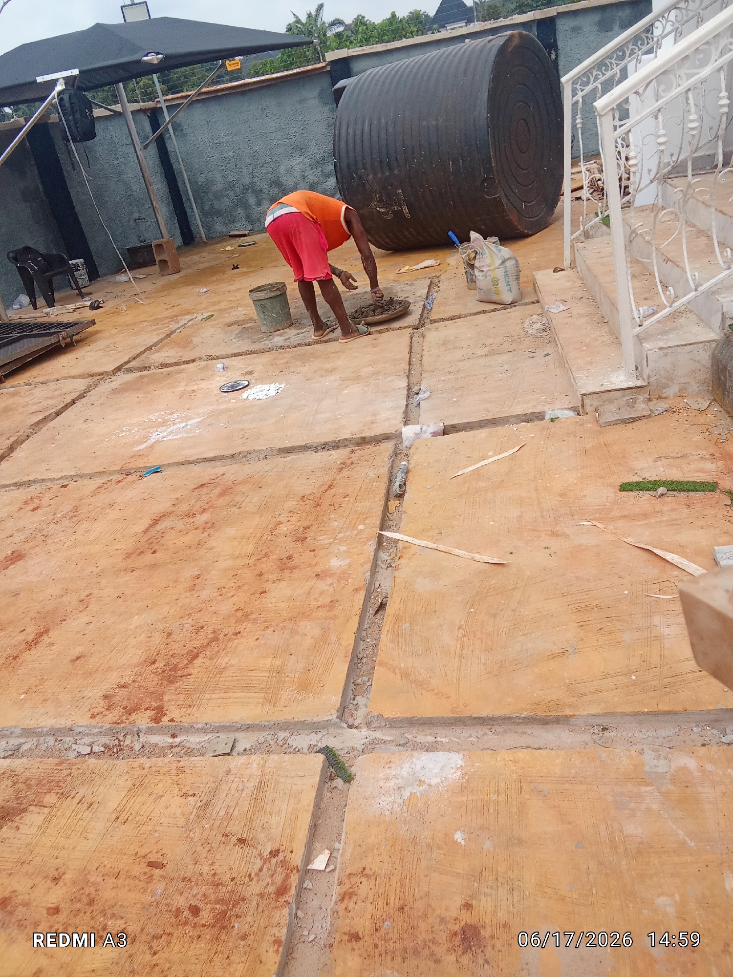 This is a medium shot of a man working outdoors in what appears to be a construction or renovation area. He is wearing an orange sleeveless shirt and red shorts, and he is bent over, seemingly mixing or applying cement. He is barefoot. The ground is paved with large, reddish-orange tiles or concrete slabs, separated by narrow trenches.
In the background, a very large, black, corrugated water tank dominates the scene. To the right, there is a white ornamental staircase with railings. A black tarp or awning is visible in the upper left, supported by metal poles. There are various construction materials scattered around, including a bag of cement, a bucket, and some debris.
The lighting suggests it is daytime, possibly late morning or afternoon. The overall atmosphere appears to be one of active work and construction.
Visible text includes "REDMI A3" in the bottom left corner and "06/17/2026 14:59" in the bottom right corner, which indicates the date and time the photo was taken. The location context is Nawfia, Nigeria.