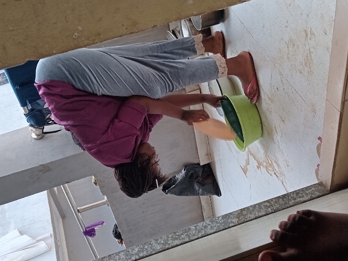 A woman in Umueshi, Nigeria, is depicted bent over a tiled floor, engaged in a cleaning or preparation task. She wears a loose purple t-shirt, light gray sweatpants with lace trim at the ankles, and pink flip-flops. Her dark, braided hair is visible. Her hands are positioned over a light green plastic basin on the floor. In her right hand, she holds an orange plastic plate, while her left hand places a long green vegetable into the basin, which contains water. Visible water is spilled on the light-colored tiled floor around the basin. In the background, a black plastic bag rests against a concrete wall. A metal railing is visible further back, draped with purple fabric, with white laundry hanging beyond. To the right, several metal containers are present. A bare foot is visible in the bottom-left foreground, positioned near a doorway frame.