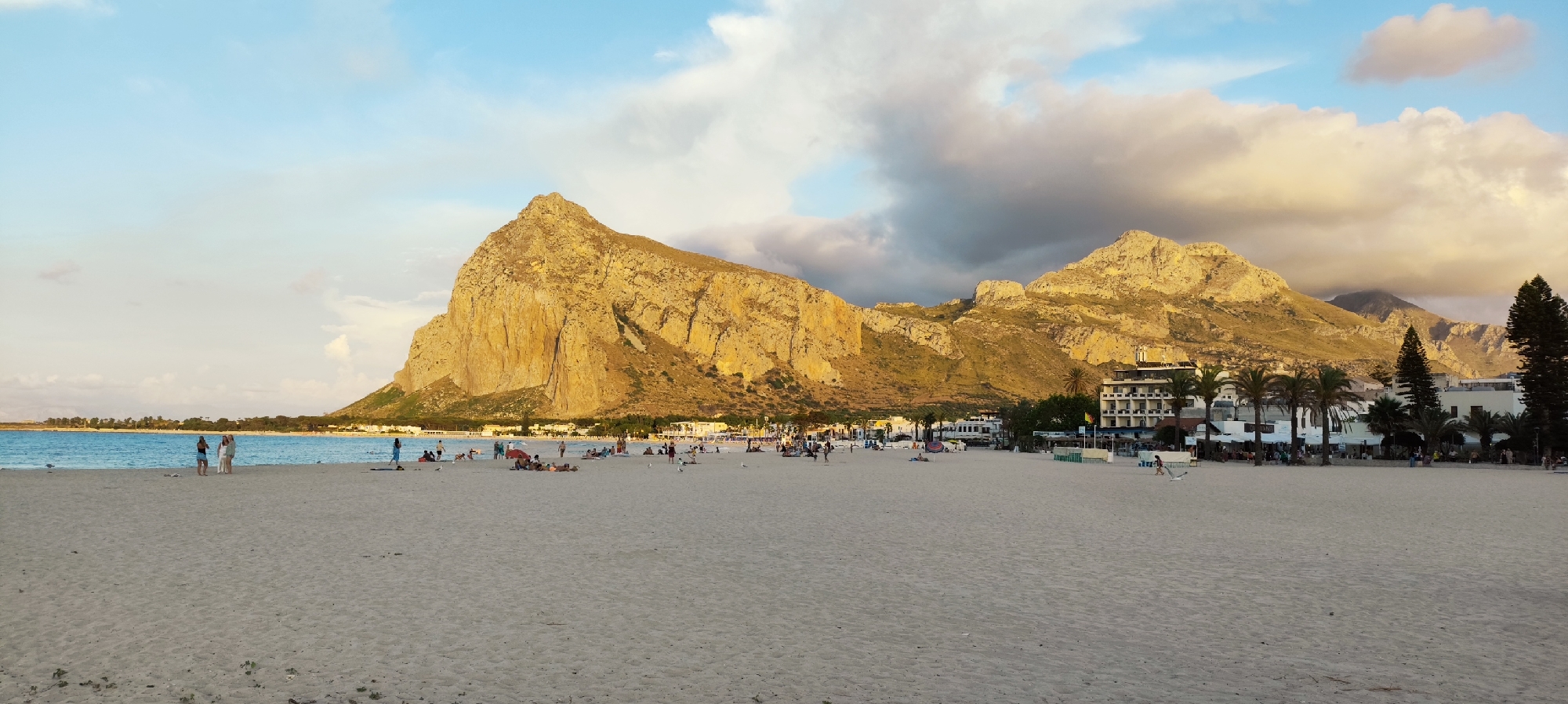 The image depicts a wide, sandy beach in San Vito Lo Capo, Italy, with a prominent rocky mountain dominating the background. The sea is a vibrant blue, meeting the shore gently. Several people are scattered across the beach, some walking, others relaxing on towels or sitting. A few individuals are standing near the water's edge. In the middle ground, a line of buildings, including a hotel with multiple stories and smaller structures, are visible behind a row of palm trees. The sky is a mix of blue and white clouds, with some areas tinged with the warm light of sunset. The overall scene is one of a peaceful, populated beach area with a dramatic natural backdrop.