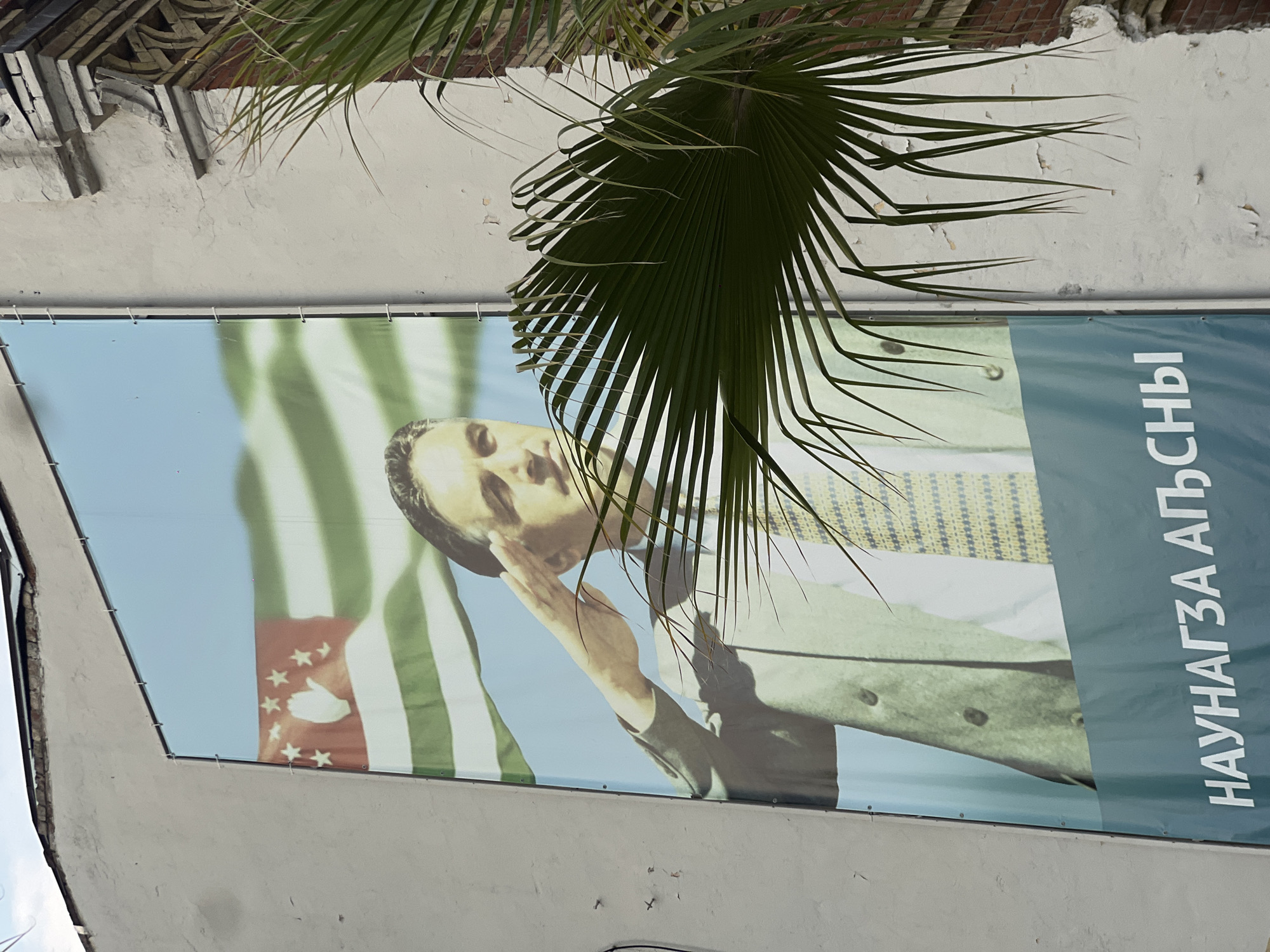 This media file depicts a large banner hung on a white, textured wall, partially obscured by the fronds of a palm tree. The banner features a portrait of a man in a suit and tie, with his hand saluting. Behind him, a flag with green and white stripes and a red canton with white stars and a hand symbol is visible. To the right of the portrait, vertical white text in an unfamiliar script is present. The overall impression is that of a political or nationalistic display, likely in a warm climate given the palm tree. The lighting suggests it could be daytime. The banner appears to be made of vinyl and is attached to the wall with visible grommets. The wall itself shows signs of age and weathering, with peeling paint and some cracks.