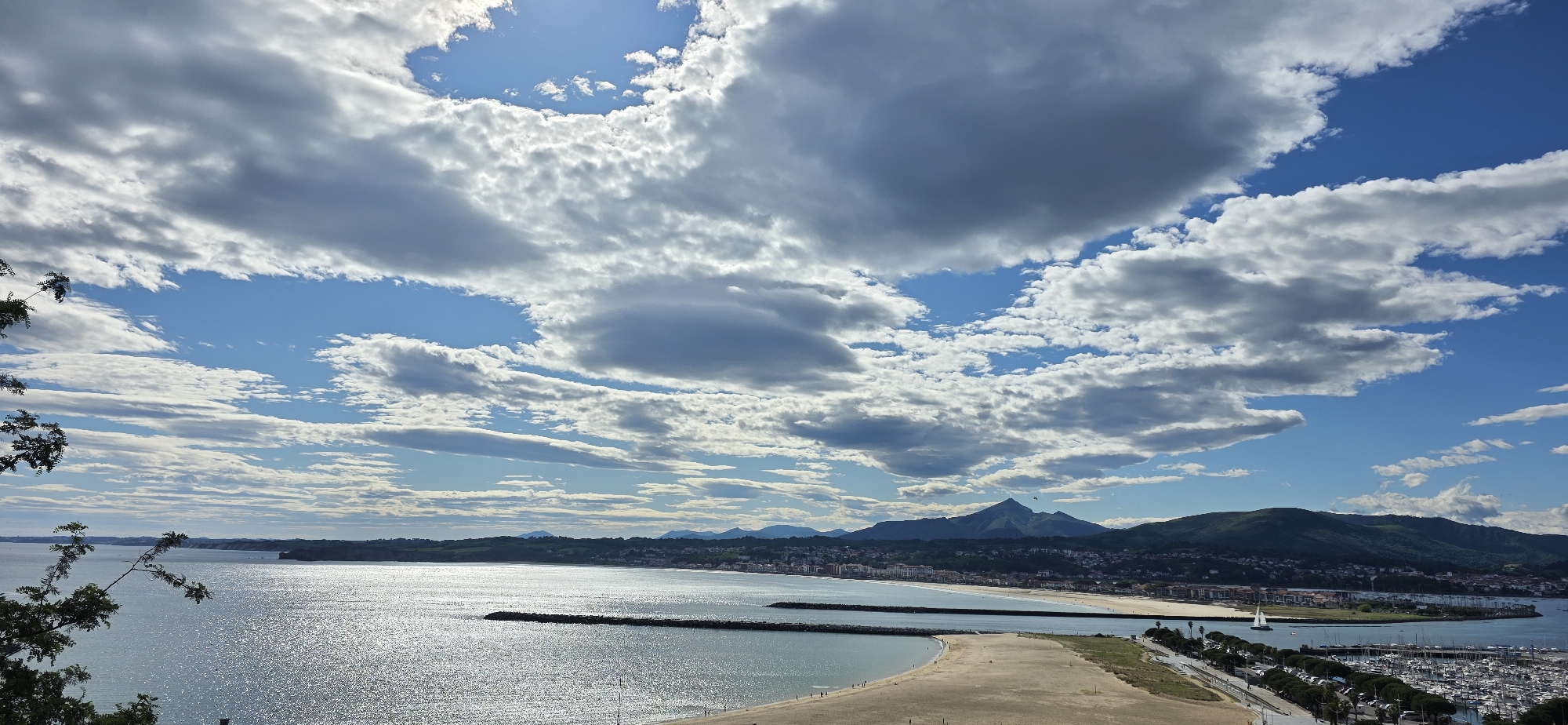 A panoramic view of Hondarribia, Spain, captured on a sunny day with dynamic clouds populating the sky. The city rests along the coastline, visible in the middle ground with buildings clustered together. To the left, the coastline extends into the distance, merging with the horizon. Mountains loom in the background, their silhouettes against the sky.
The foreground features the ocean, reflecting sunlight and creating a sparkling effect. A sandy beach and a breakwater are visible. A road runs parallel to the marina, lined with trees and possibly parked vehicles. The marina is filled with boats. A sailboat glides on the water near the shoreline. People are walking on the beach and along the breakwater. The photograph is taken from an elevated viewpoint, likely a hill or cliff, offering a comprehensive view of the landscape. Some foliage in the far left hints to the viewpoint position.