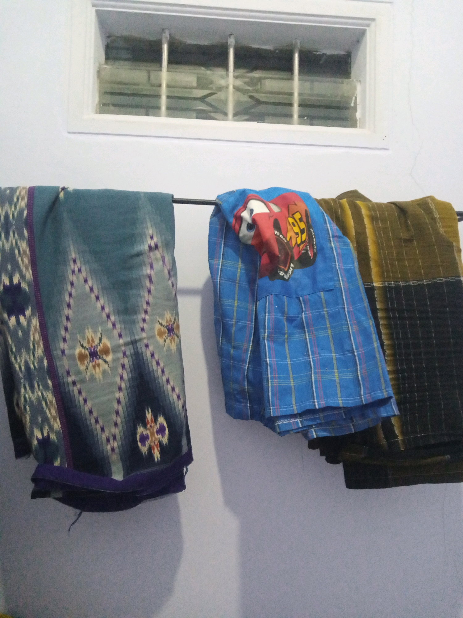 The image depicts three pieces of fabric hanging on a clothes rack in front of a white wall. The fabric on the left is teal with a diamond-shaped pattern in various colors including purple, brown, and white. The middle fabric is blue with a plaid pattern and features a large graphic of Lightning McQueen, a character from the Disney Pixar movie "Cars", on it. The fabric on the right is a darker color, possibly brown and black, with vertical stripes. Above the fabrics, there is a rectangular window with horizontal bars across it. The scene appears to be indoors, possibly in a home, given the presence of a clothes rack and window. The lighting suggests it is daytime, but the interior setting implies artificial light might also be present or that the area is dimly lit. The overall impression is that of laundry being dried indoors. The location is specified as Sumbergirang, Indonesia.