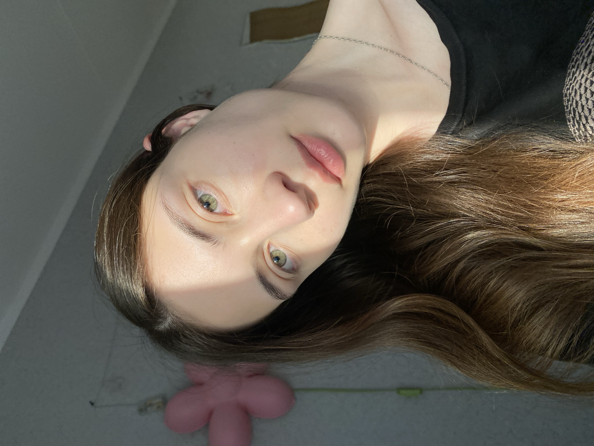 A medium-shot, close-up photo of a fair-skinned woman with light brown hair and green eyes, oriented so she is almost upside down. She is wearing a thin silver chain necklace and a black garment. In the lower left corner is a pink, flower-shaped object. The background appears to be a gray floor and wall. There is no verifiable information indicating that the photo was taken in Votkinsk, Russia.
