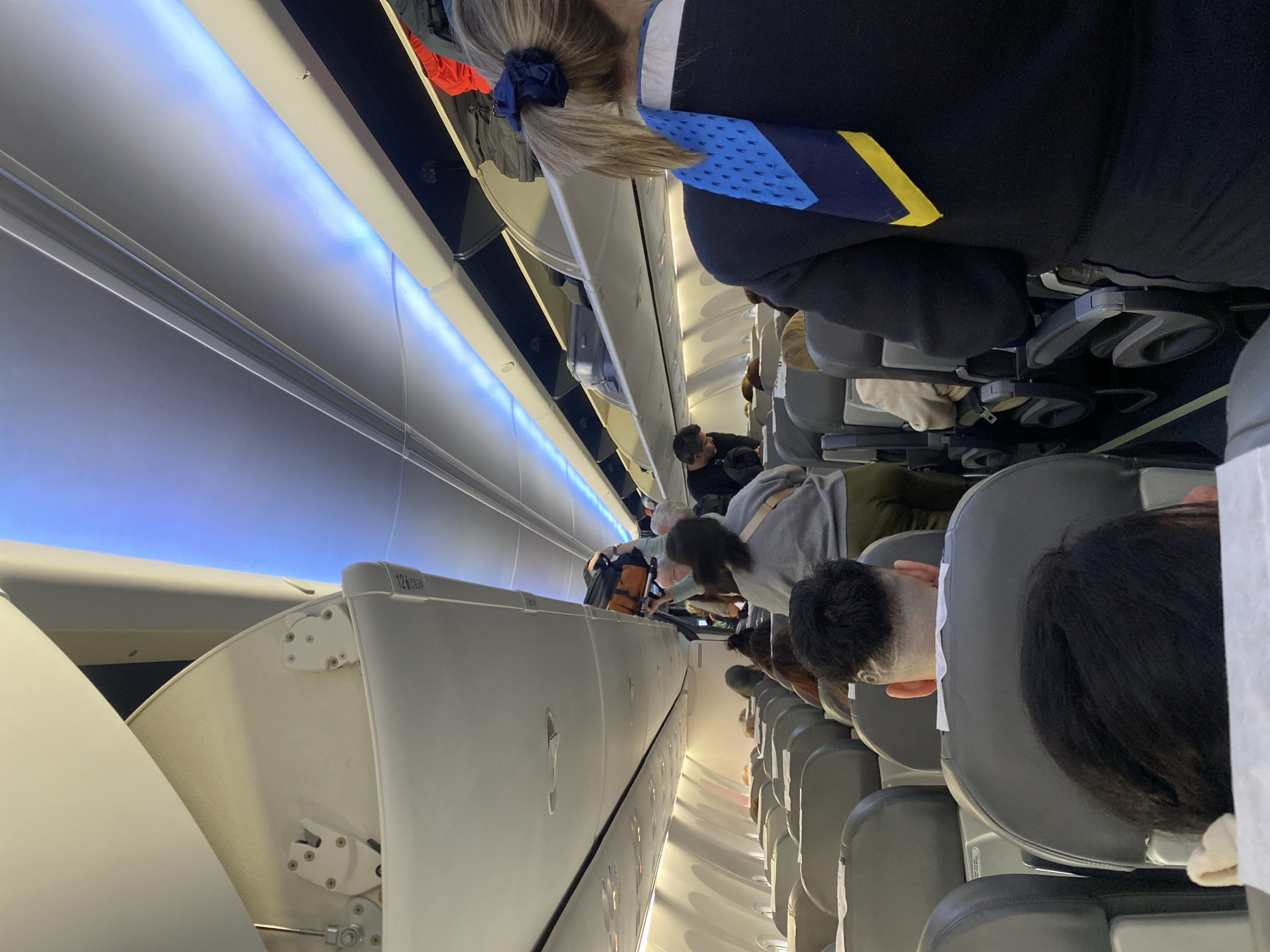 The image captures the interior of an airplane cabin, oriented on its side, showing the aisle running horizontally across the frame with rows of seats extending vertically. The scene appears to be during boarding or deplaning, given the presence of standing passengers and open overhead bins. The context provided indicates this scene is in Ushuaia, Argentina.
Numerous passengers are visible, mostly from behind. In the immediate foreground, the back of a person with light brown hair tied in a ponytail with a blue scrunchie is prominent. They are wearing a dark top and a distinctive blue and yellow patterned scarf or tie. Further down the aisle, other passengers are seated, their heads resting against headrests. One individual has a short, shaved haircut with a visible pattern on the side of their head.
The overhead bins are a key feature, some of which are open. The text "12A" and "12B" is visible on the edge of an open bin, indicating seat numbers. The cabin interior features modern design elements, including a strip of blue accent lighting running along the ceiling, casting a glow on the white panels of the overhead compartments. The overall atmosphere is that of a typical commercial flight.