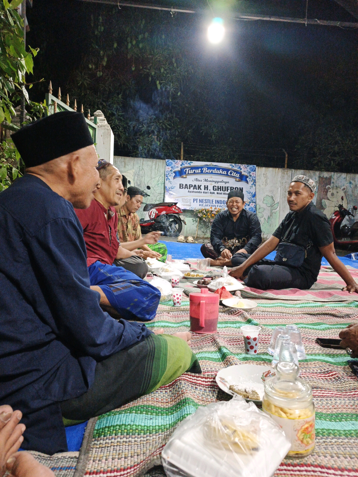 This is a nighttime, medium shot photograph taken outdoors in Sumber Gedang, Indonesia. The scene depicts a group of men sitting on a woven mat on the ground, engaged in a gathering. A banner in the background reads "Turut Berduka Cita" which translates to "Condolences" and mentions "BAPAK H. CHUFRON". The presence of the banner indicates a memorial or condolence gathering.
In the foreground, a man in a dark blue shirt and a black peci (cap) is seen from the side, looking to the left. He is wearing a green and black sarong. To his right, other men are seated, some in traditional attire, including pecis and sarongs. One man in the center wears a red shirt and blue sarong. Another man to his right wears a patterned shirt. Further to the right, a man in a black shirt and pants, wearing a patterned peci, is looking towards the camera and smiling. He has a black sling bag across his chest.
On the mat, various objects are spread out. There are plates with food, cups, and a large red pitcher. A glass jar filled with what appears to be crackers or biscuits is also visible. In the background, a red motorcycle is parked, and there is a wall with some artwork or decoration. Smoke is visible rising from the motorcycle area. The lighting is provided by an overhead bulb, casting a warm glow on the scene. The overall atmosphere suggests a somber yet communal event.