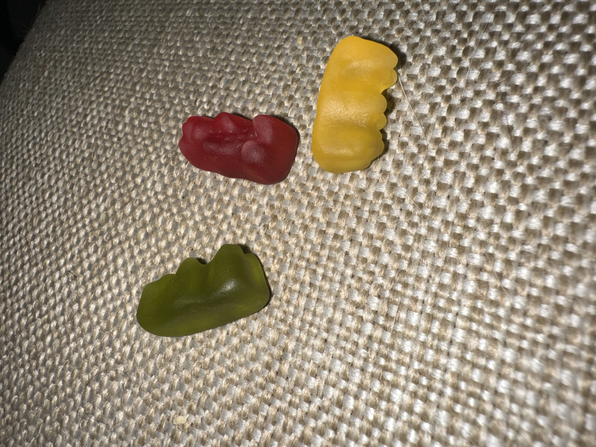 The image shows three gummy candies - red, yellow, and green - on a tan woven surface. No location information is discernible from the image.