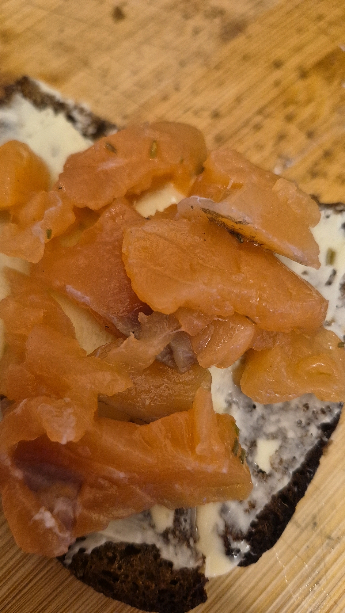 A close-up, overhead shot captures smoked salmon on dark bread with butter on a wooden surface. The salmon, a vibrant orange, is thinly sliced and arranged in a pile, some pieces slightly curled. A creamy white spread, likely butter, is visible beneath the salmon and on the dark, dense bread, which appears to be a slice of rye. The wooden surface has a distinct grain pattern and is oriented diagonally. No people or specific urban details indicating the city of Krauja, Latvia, are present.