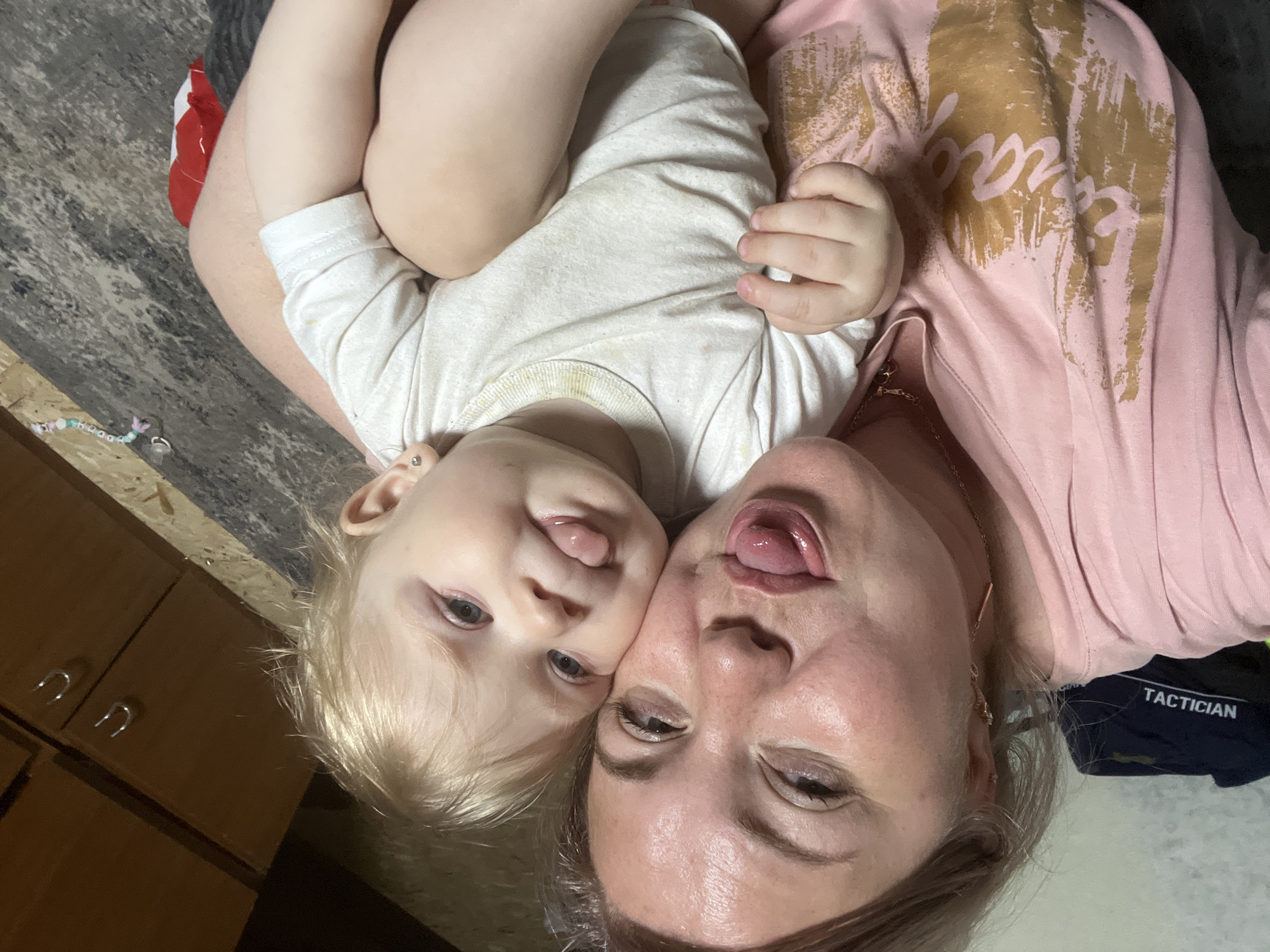 An extreme close-up selfie shows a Caucasian woman and a baby. The woman is on the left, with her head tilted and tongue sticking out. She has blonde hair and is wearing a pink t-shirt with a gold design. The baby is on the right, with its head also tilted and tongue sticking out, facing the woman. The baby has blonde hair and is wearing a white onesie. They are both positioned very close to the camera, filling most of the frame. The background appears to be an indoor setting with wooden cabinets and a patterned floor. There is no information about the location in the city of Фоки, Russia.