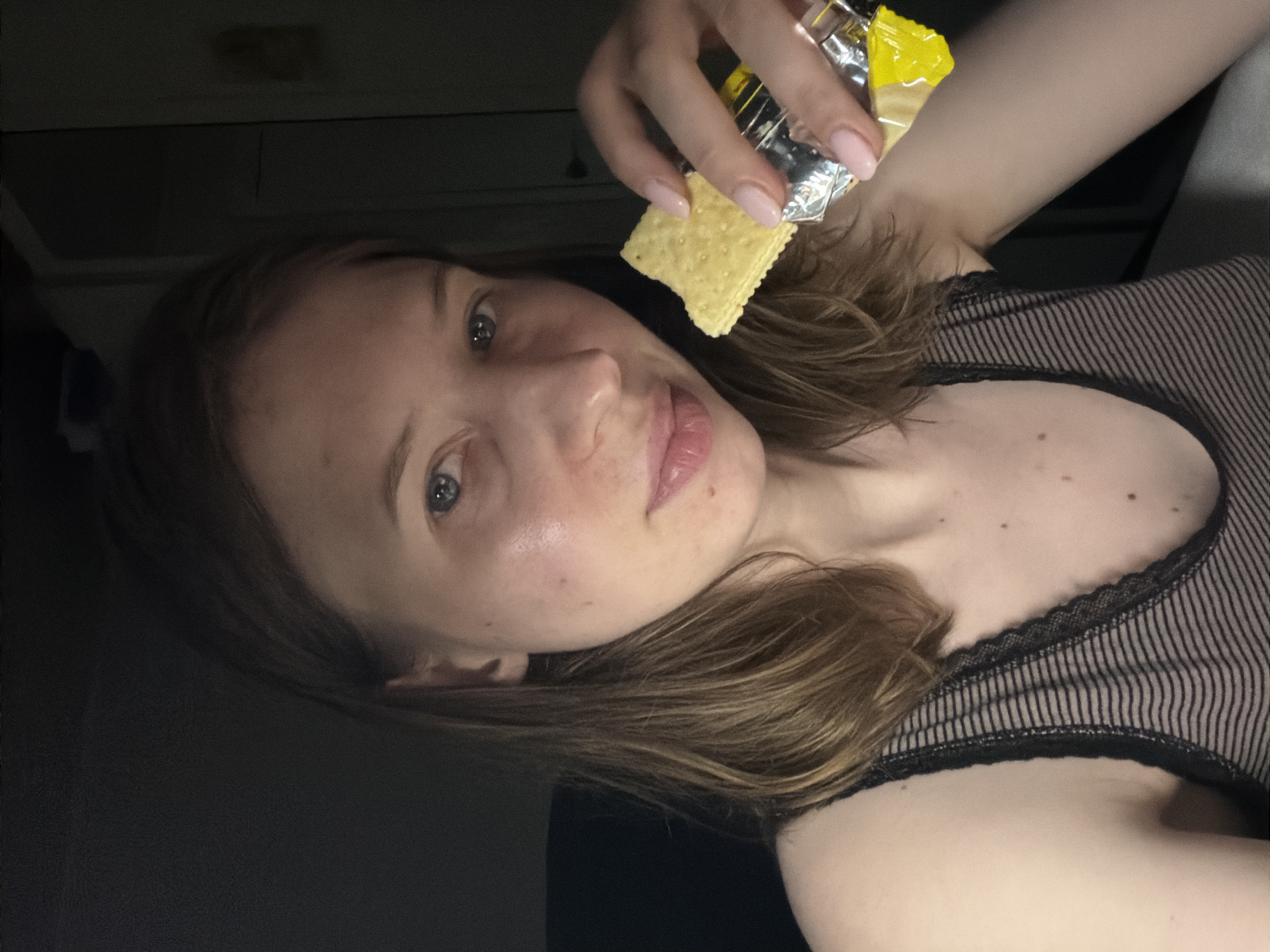 A young adult female is pictured in a dimly lit indoor setting. She has shoulder-length brown hair and is looking towards the camera with blue eyes. Her skin appears to have some redness, particularly on her cheeks. She is wearing a black and grey striped tank top with lace trim. In her right hand, she is holding a cracker that appears to be from a yellow and silver wrapper. The background is dark and indistinct, with hints of furniture or a wall. The scene does not provide any identifiable landmarks or contextual information to suggest a specific location within Dongducheon-si, South Korea.