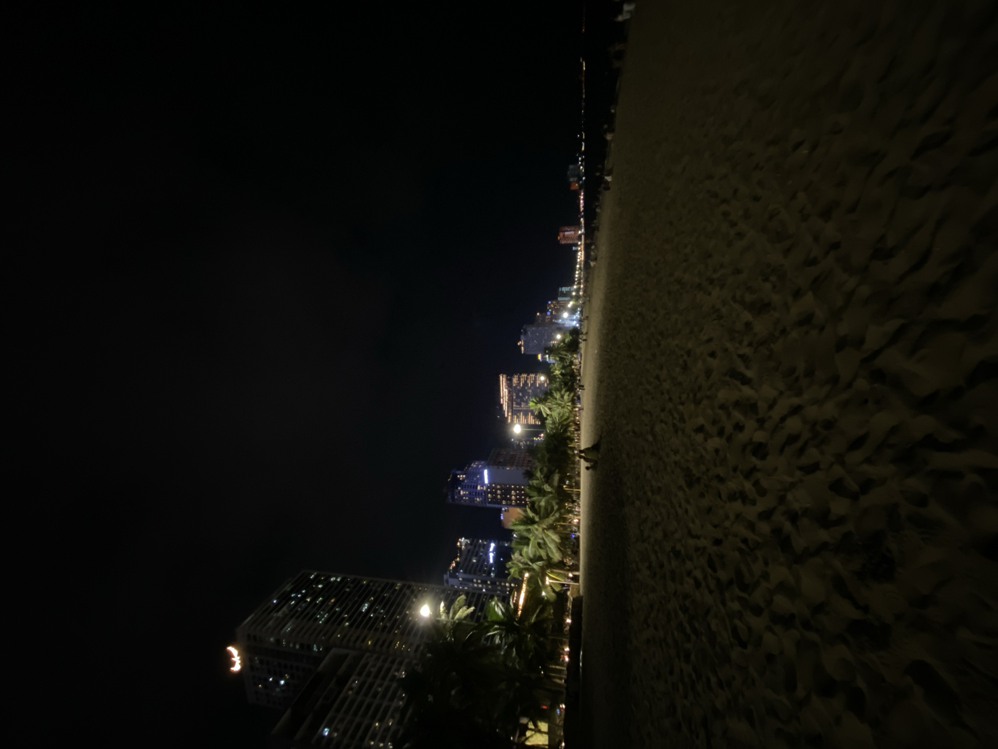 The image depicts a nighttime view of a sandy beach with a city skyline in the background. The perspective is from ground level on the beach, looking towards a line of buildings illuminated by artificial lights. The sand in the foreground is textured, showing footprints and ripples, suggesting it is a well-used beach. The buildings in the distance vary in height and architectural style, with many of them featuring numerous lit windows, indicating a vibrant urban area. Palm trees are visible along the edge of the beach, bordering the city. The overall scene is dark, typical of nighttime, with the city lights providing the main illumination.
There appear to be a few indistinct figures walking on the beach and near the treeline. The atmosphere seems peaceful and quiet, given the absence of significant activity. The location is identified as Nha Trang Ward, Vietnam, which is a coastal city known for its beaches. The time of day is clearly night, and there are no visible indicators of weather conditions, but the darkness suggests a clear sky or at least no significant precipitation. The mood is serene, with the contrast between the dark natural elements and the bright man-made city lights creating a visually appealing composition. No visible text is present in the image.
