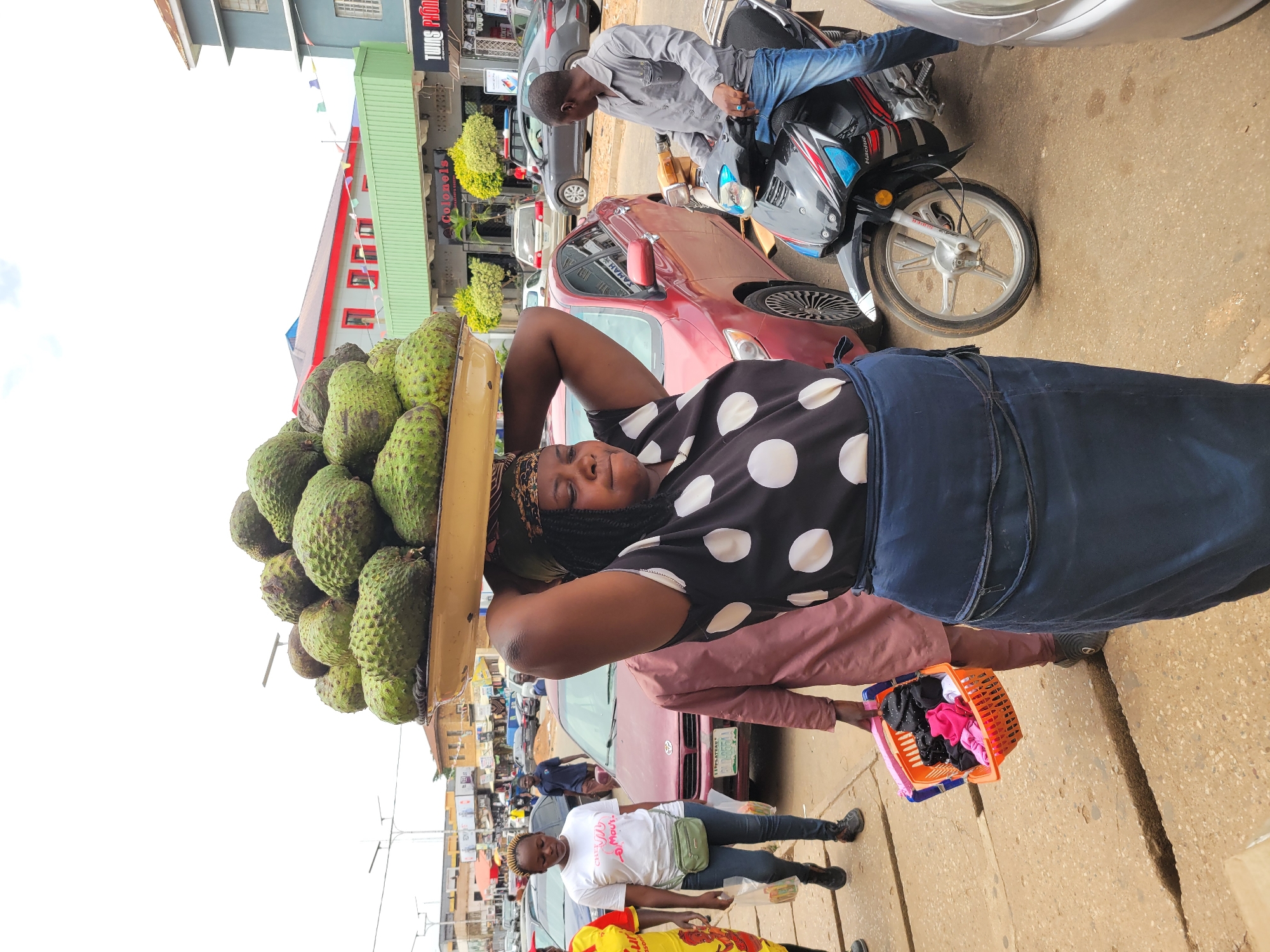 The image captures a street scene in Jos, Nigeria. A woman stands in the foreground, balancing a tray laden with numerous soursop fruits on her head. She wears a black and white polka-dot top and a dark blue skirt. Her hands are placed behind her head, seemingly supporting the tray.
Behind her, several people are visible, including a man sitting on a motorcycle. A maroon car is parked nearby. Other people are walking on the street. One person is holding a colorful plastic basket.
The background reveals urban buildings, suggesting a commercial area. The image indicates a typical scene of street vending within a city environment.