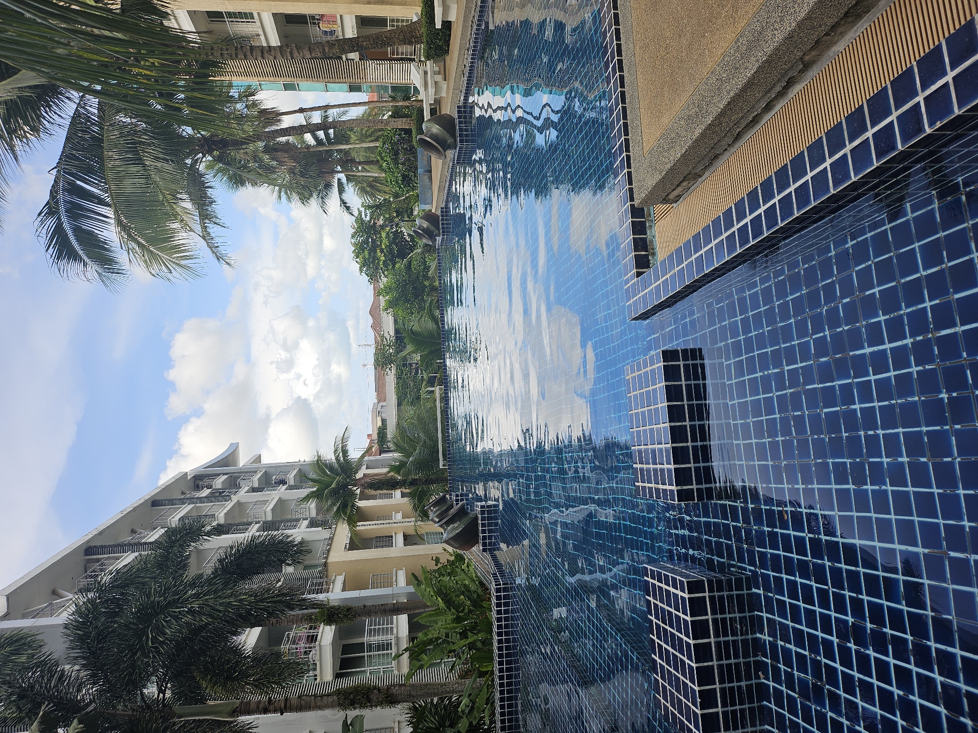 A high-angle, medium shot captures a swimming pool at the Patong Bay Garden Resort in Kathu, Thailand. The resort's white building with multiple balconies is visible on the left and right sides of the frame, partially obscured by lush palm trees. The swimming pool itself is tiled with small, dark blue squares, and its surface reflects the cloudy blue sky and surrounding greenery. Two large, dark, urn-shaped decorative objects are placed at the edge of the pool. The pool area features a walkway with a sandy-colored surface and a drainage grate. A few white lounge chairs are visible on the right side of the walkway. The image is taken during the day, with bright natural light.