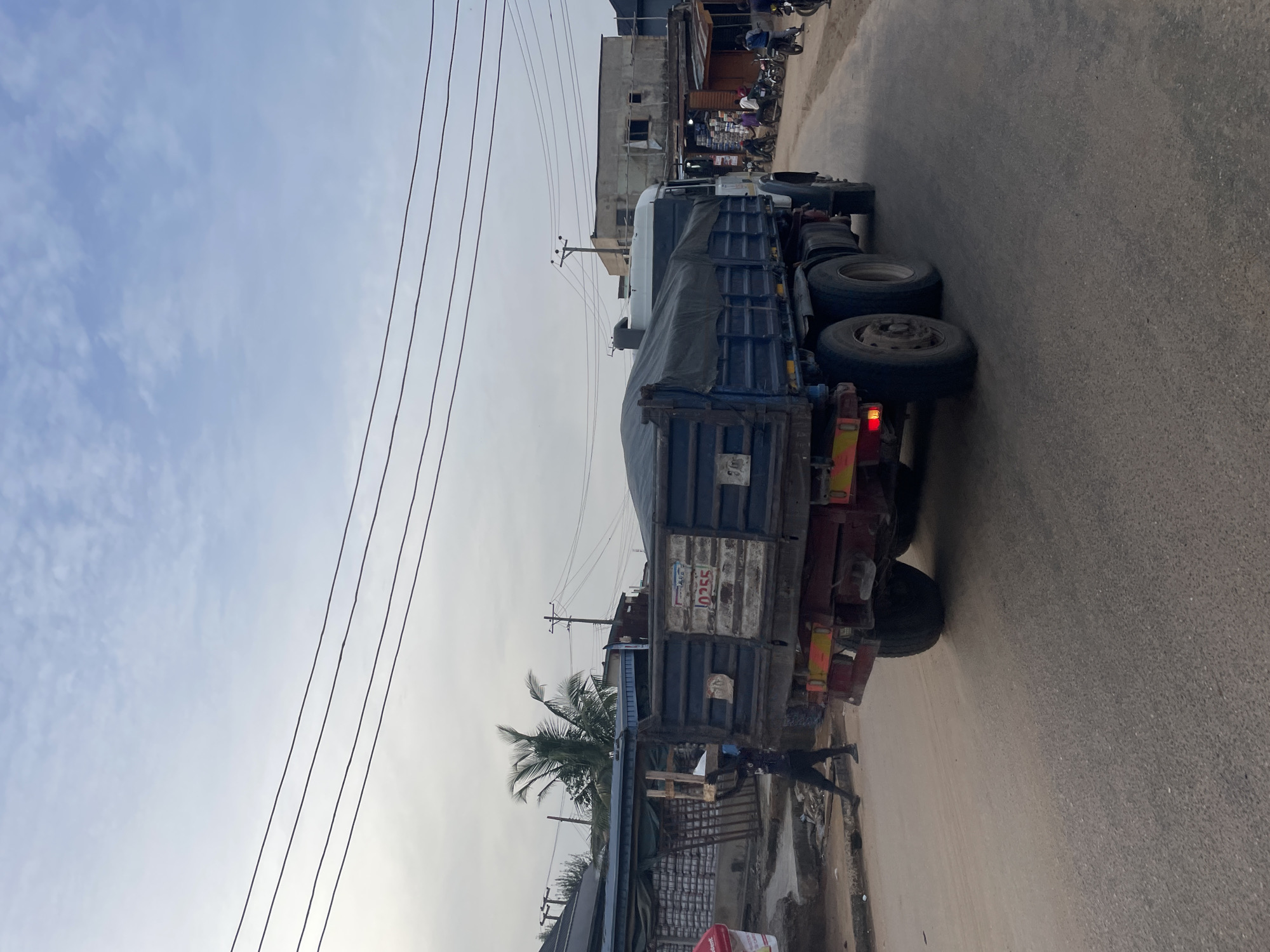 A photograph depicts a truck on a road in an urban setting, the city is unknown. The truck has a blue container with a white cover. There are visible power lines overhead. The background features buildings and motorcycles, suggesting a commercial or residential area. A person is seen near the truck. The image appears to be taken during the day under a cloudy sky.