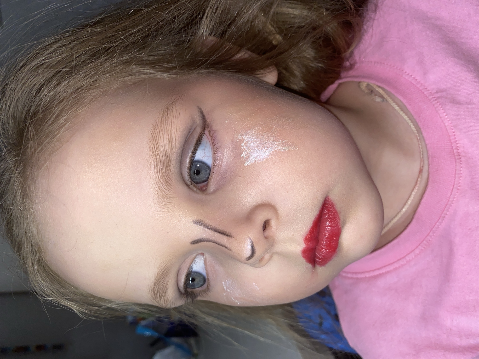 A close-up, slightly rotated view of a young girl with light hair and blue eyes is shown. The girl is wearing a pink shirt. The girl has makeup applied; red lipstick, white cream on her cheek, and a black line drawn down her nose. The background appears to be indoors, with a blue and white object visible out of focus. It is not possible to identify the location as Votkinsk, Russia based solely on this image.