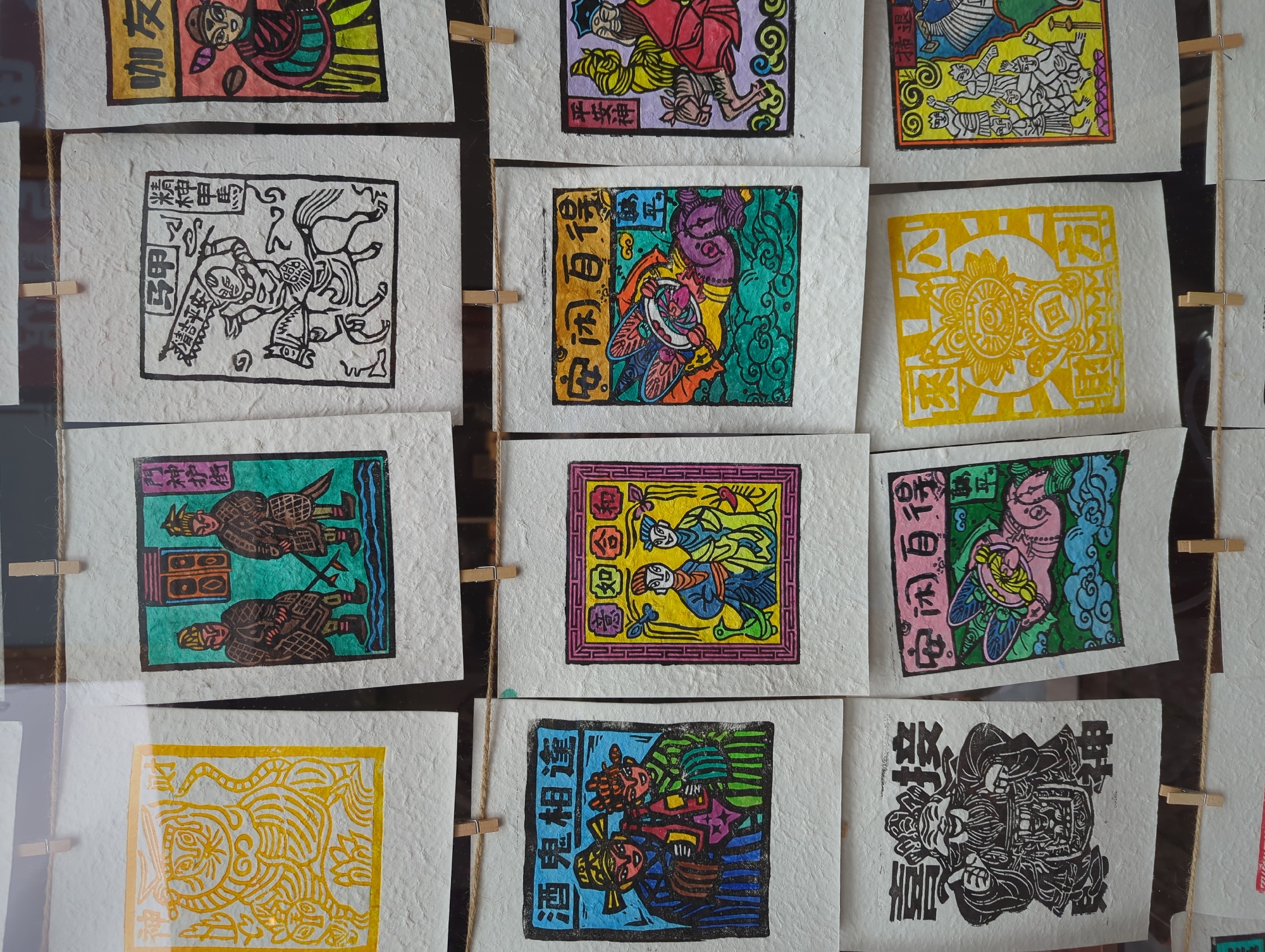 This image depicts a collection of traditional Chinese paper-cut art prints, likely displayed for sale in Gucheng District, China. The prints are hung on a string using clothespins, suggesting an outdoor or market stall setting. Each print is rectangular and features colorful, stylized depictions of figures, animals, and symbolic imagery, along with Chinese calligraphy. Some prints show historical or mythological characters, while others display auspicious symbols. The artwork is displayed on textured, off-white paper. The background is blurred, indicating a shallow depth of field, with glimpses of other similar artworks and possibly the environment of the market.