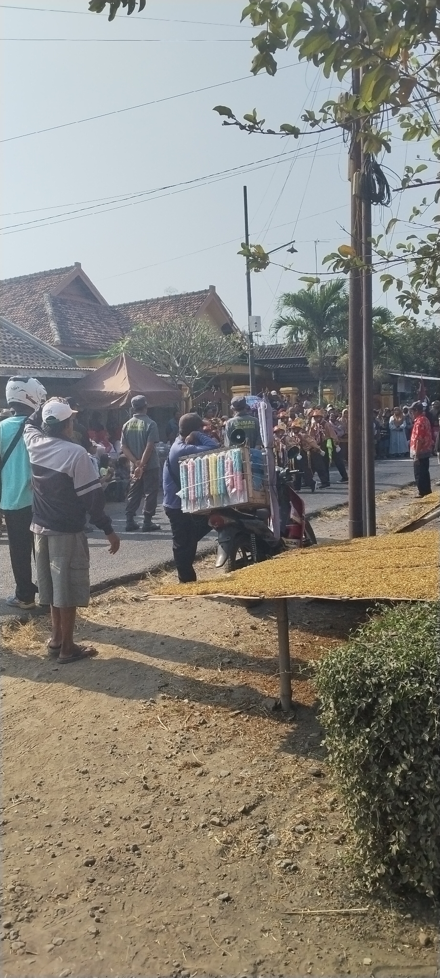 In Yosowilangun Kidul, Indonesia, on a bright, clear day, a street scene unfolds with a gathering of people. In the foreground, a person wearing a blue shirt and shorts stands near a table laden with a dry, granular material, possibly grains. Next to them, a motorcycle is parked, topped with a rack carrying a colorful assortment of cylindrical items, appearing to be snacks or packaged goods. Behind this, a group of individuals, some in uniform, are observing or participating in an event. Further back, buildings with traditional tiled roofs line the street, and a dense crowd is visible, suggesting a local event or gathering. The environment is dusty and dry, with sparse vegetation and utility poles with hanging wires suggesting an urban or semi-urban setting.