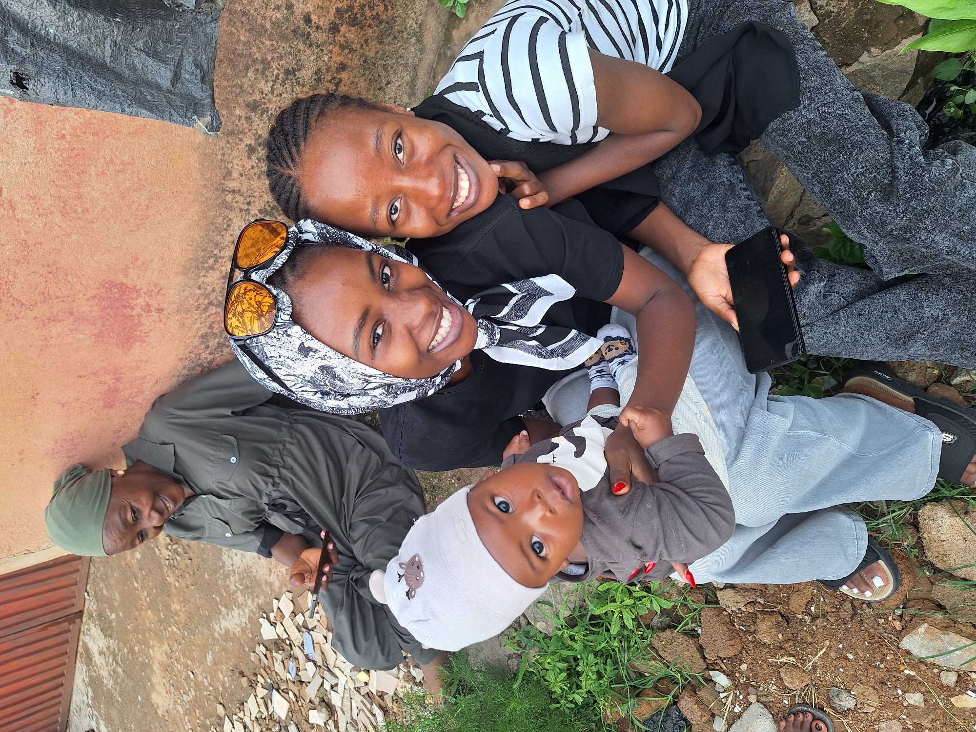 Four individuals are gathered outdoors in Sabon Tasha, Nigeria, against an orangish-brown wall. An elderly woman in the background wears an olive green garment and headwrap, smiling down at a dark smartphone in her right hand. To her right, two younger women are seated in the foreground, facing the camera with smiles. The woman on the left wears a black t-shirt, a black and white patterned headscarf, and orange-tinted sunglasses on her head. She holds an infant, dressed in a grey beanie with a deer emblem and a grey top, who looks forward. Her left arm is around the second young woman. The second young woman wears a black and white striped t-shirt over a black garment and grey acid-wash jeans, holding a dark smartphone in her left hand. The ground consists of bare earth with scattered light-colored debris and sparse green vegetation. A dark, tattered fabric is partially visible against the wall on the right.
