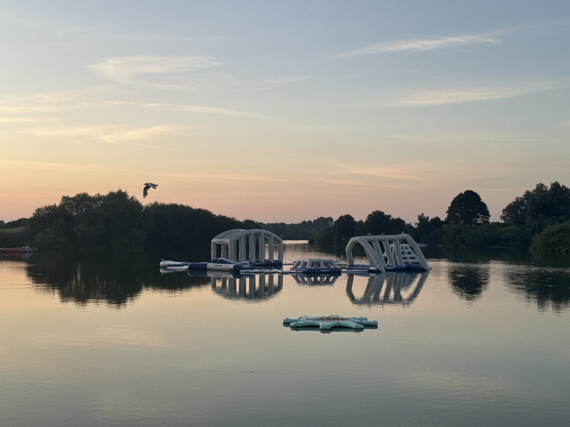 This image depicts an outdoor scene at the Craigavon, United Kingdom, likely a lake or reservoir, during twilight. The sky is a gradient of soft blues and pinks, indicative of sunset or sunrise. A single bird is in flight in the upper left portion of the frame.
The water's surface is calm, reflecting the sky and the surrounding elements. In the water, there are several inflatable structures, appearing to be part of a water park. These structures are white and blue and have various shapes, including arches and ramps. One smaller, light blue inflatable is floating in the foreground.
Along the far bank of the water, a line of dense green trees forms a natural boundary. The reflections of these trees are visible on the water's surface. There are also some other watercraft, possibly kayaks or small boats, visible on the far left edge of the image, suggesting recreational activity. The overall impression is one of a peaceful, natural setting enhanced by recreational amenities.