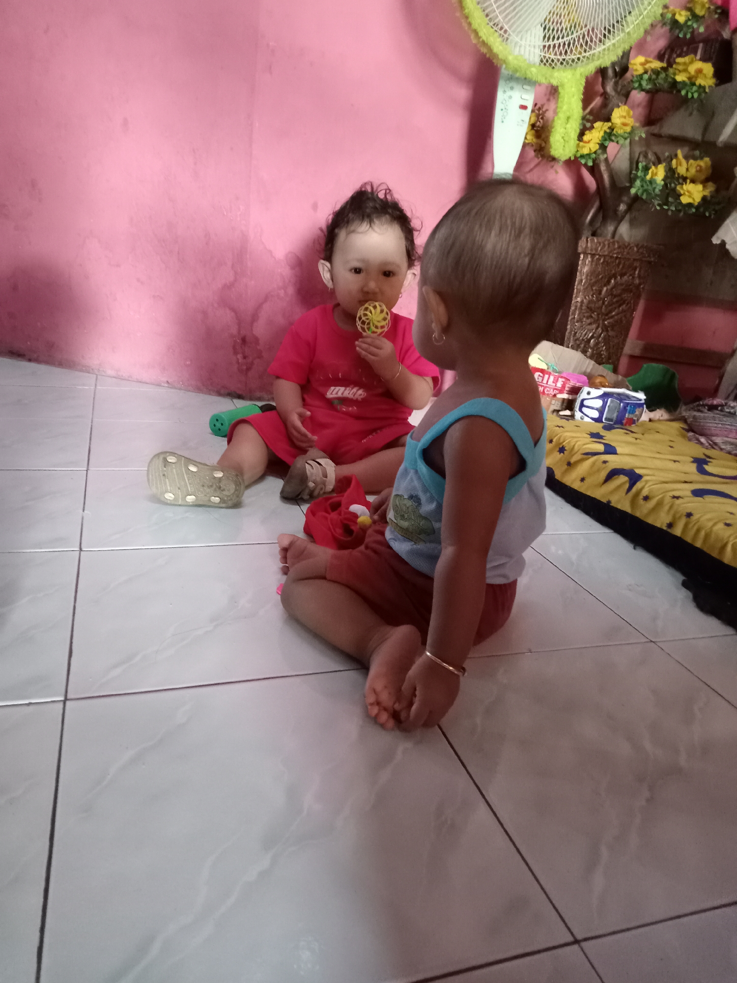 Two young children are sitting on a tiled floor in Bungur, Indonesia. One child is seated facing the camera, wearing a red shirt and holding a yellow object in their mouth. The other child is facing away from the camera, wearing a blue and white tank top and red shorts. The room has pink walls and a standing fan in the background. A yellow and blue patterned mattress is also visible. There are toys scattered around the children.